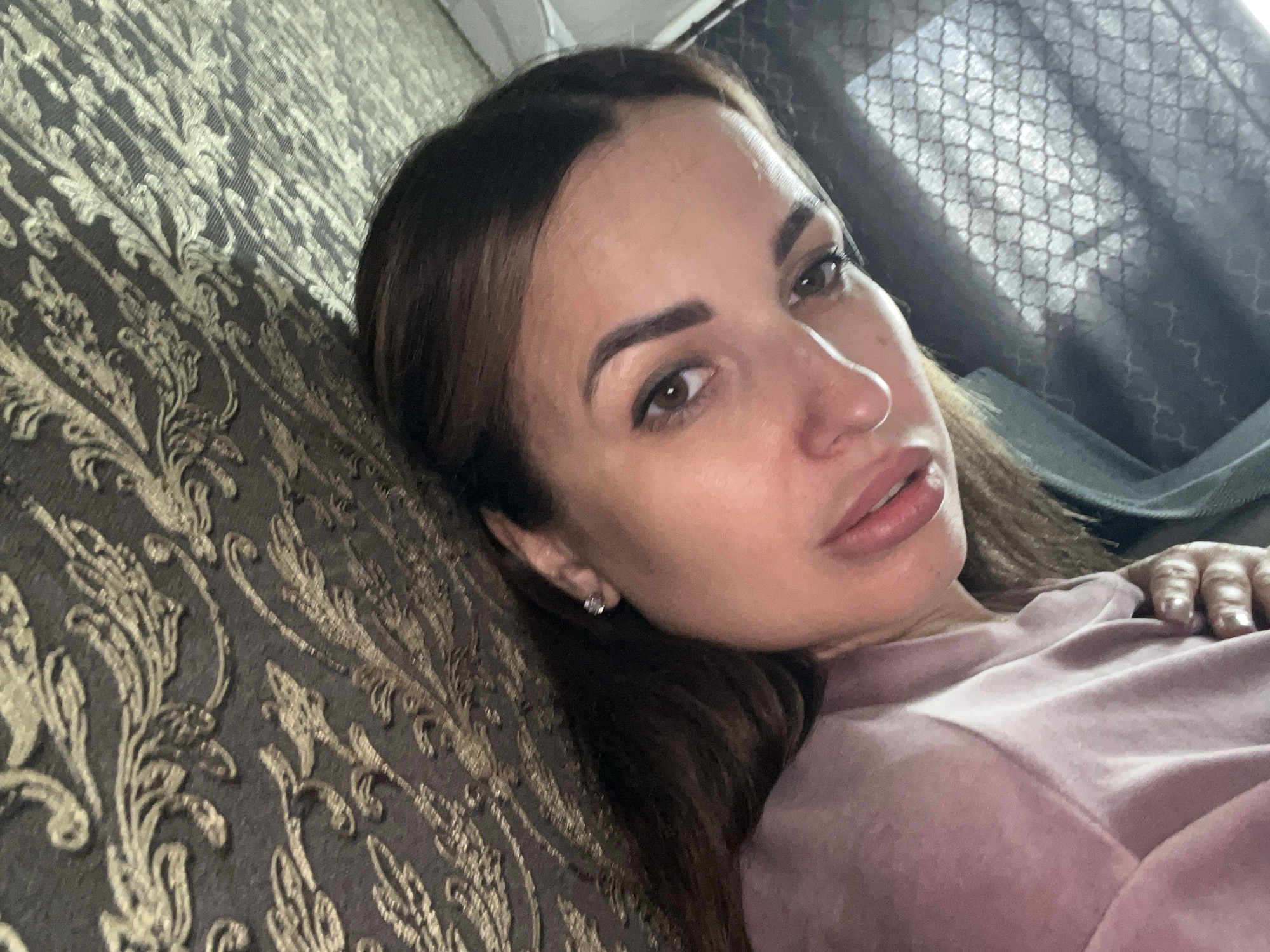 The image shows a close-up, angled selfie of a woman with brown hair and brown eyes, looking at the camera. She is wearing a light pink top and has a nose ring and stud earrings. She is lying down, with her head resting on a textured surface that appears to be upholstery with a gold patterned design. A portion of a black textured material is visible behind her. The lighting suggests an indoor environment. The image does not provide any information about a specific city or location.