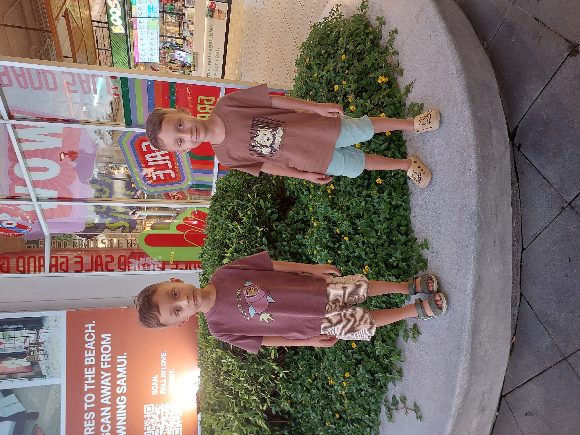This is a vertical, eye-level, medium shot of two young boys standing in front of lush green foliage and a store window display. Both boys appear to be in their early to mid-childhood. The boy on the left, closer to the camera and store window, is wearing a brown t-shirt with a graphic of a dog and text that appears to say "DARE YOU" and light blue shorts. The boy on the right, further back, is wearing a reddish-brown t-shirt with a rocket graphic and the text "ROCKET TIME", and striped shorts. Both boys are wearing light-colored sandals.
The background shows a brightly lit store interior with vibrant signage, including what appears to be sale advertisements with colorful stripes and bold text such as "WOW" and "SALE DI". The text on the store windows is partially obscured and reflected, but some words like "fruit. real vegetables. de for real." and "ADD GRAND SALE GR" are legible. Another sign on the far left, near the boy on the left, has text in English and appears to be an advertisement for "BAAN CHAWENG, SAMUI" with a QR code and the text "SCAN. FALL IN LOVE. INVEST." This strongly suggests the location is Baan Chaweng, Thailand.
The boys' expressions are neutral, and the overall mood is casual and bright, typical of a sunny day in a commercial area. The lighting suggests it is daytime. The scene captures a moment of children posing in front of a retail environment.