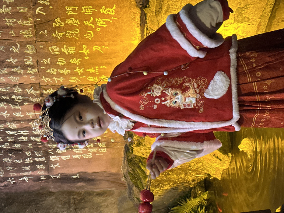 An Asian female child, estimated 6-8 years old, is centered in a culturally themed area within Yuzhong District, China. She wears a traditional red Chinese jacket with white faux fur trim, adorned with elaborate gold embroidery depicting a stylized mythological creature holding a scroll, along with a patterned red skirt. Her head is crowned with a gold-colored, ornate headpiece featuring red spheres, blue beads, and dangling decorative elements. Her face has minimal makeup, including glitter on the cheeks and a small red dot on her forehead. She holds a wooden skewer with three candied hawthorns (tanghulu) in her outstretched right hand, looking directly at the viewer with a neutral expression.
The background consists of a large, textured stone wall intensely illuminated by warm yellow light, displaying dense Chinese calligraphy carved or projected onto its surface. Below the wall, a body of water reflects the yellow light, showing faint reddish-orange fish and dark green foliage along its left edge. The overall lighting is dim, emphasizing the illuminated wall and water.