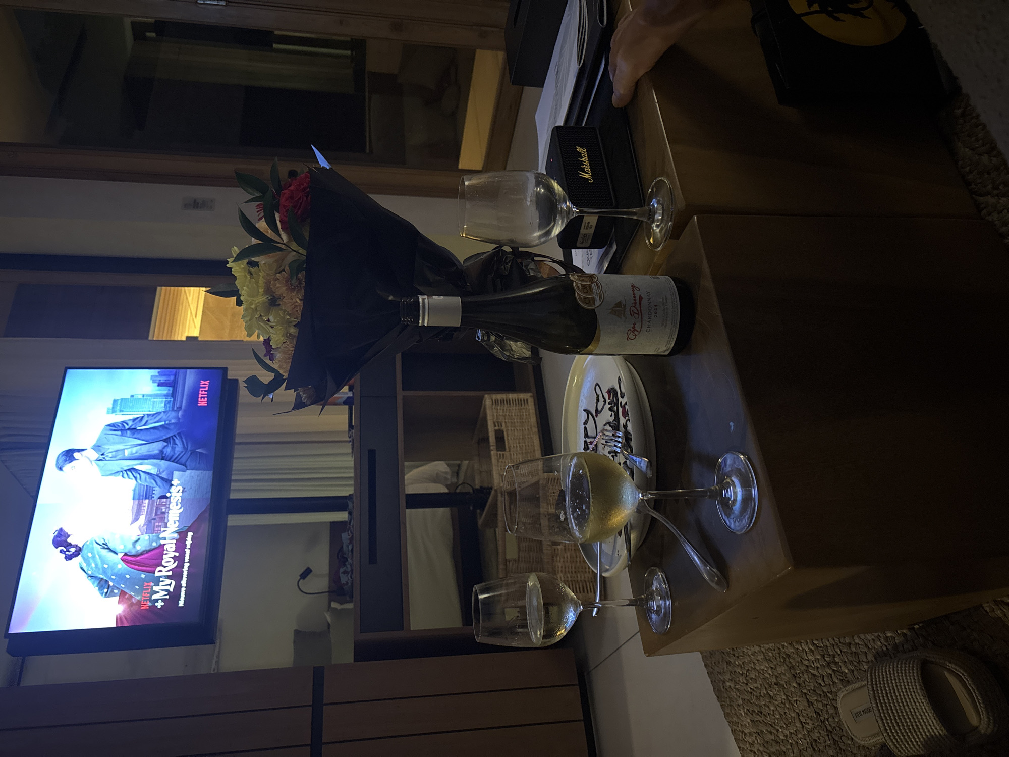 Here is a description of the image:
The image shows a table setting with a bottle of "Cape Discovery Chardonnay 2024" alongside a wrapped bouquet of flowers, indicating a celebration of some kind. There are several wine glasses, one appears to contain white wine. A slice of cake is on a plate. A Marshall speaker is also visible on the table.
In the background, a television displays the Netflix logo and a show titled "+My Royal Nemesis+". The room is dimly lit, suggesting an evening setting.
At the bottom of the frame, a pair of "Steve Madden" sandals is visible.
There is no indication that this image was taken in Kuta Selatan, Indonesia.