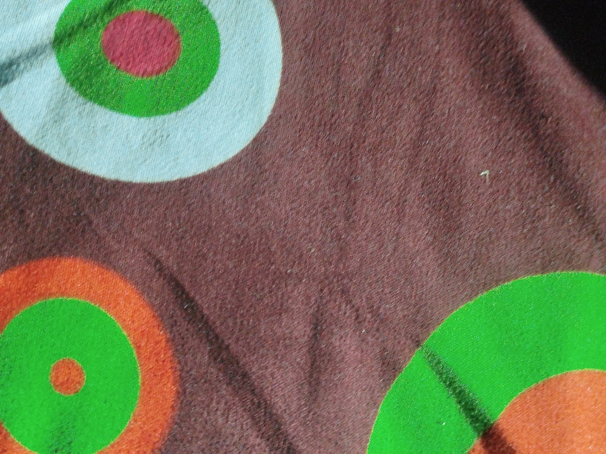 The image shows a close-up view of a brown, textured fabric with a pattern of colorful circles. There are at least three visible circular targets. One target, on the left, is light blue with a green ring and a red center. Another target, partially visible at the bottom left, is orange with a green ring and a green center. A third target, partially visible at the bottom right, is green. The fabric appears to be a soft material, possibly cotton or a blend, with a slightly fuzzy texture. The lighting suggests it might be indoors or in an area with diffused light, casting subtle shadows. There are no people or any identifiable landmarks that would allow for the determination of its location in Mansehra, Pakistan.