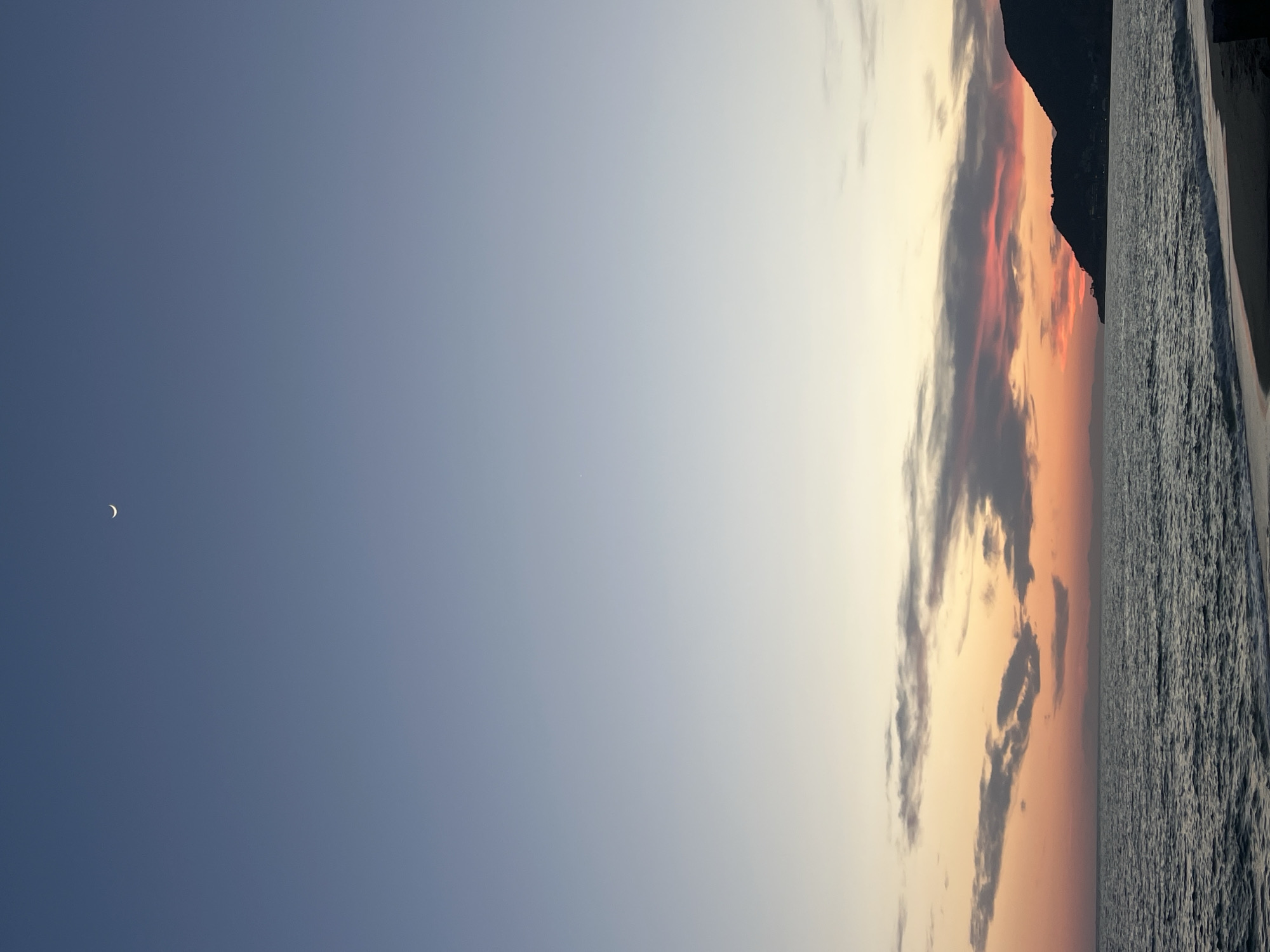 The image displays a panoramic coastal landscape at twilight, likely either sunrise or sunset, in Alanya, Turkey. The vast sky dominates the upper portion of the frame, transitioning dramatically from a deep, clear blue on the far left to softer blues, then warm yellows, oranges, and reds towards the right horizon. A delicate, luminous crescent moon is visible in the deep blue expanse of the upper left sky, indicating the twilight hours. Scattered clouds, primarily in the middle and right sections of the sky, are illuminated with hues of grey, purple, and fiery red by the setting or rising sun. The lower right portion of the image features a body of water, presumably the Mediterranean Sea, with its surface showing subtle ripples and textures. Along the horizon on the far right, a dark, undulating silhouette of a coastline or mountain range is visible, creating a stark contrast against the vibrant sky. The scene evokes a sense of tranquility and natural beauty. No discernible text is present in the image.