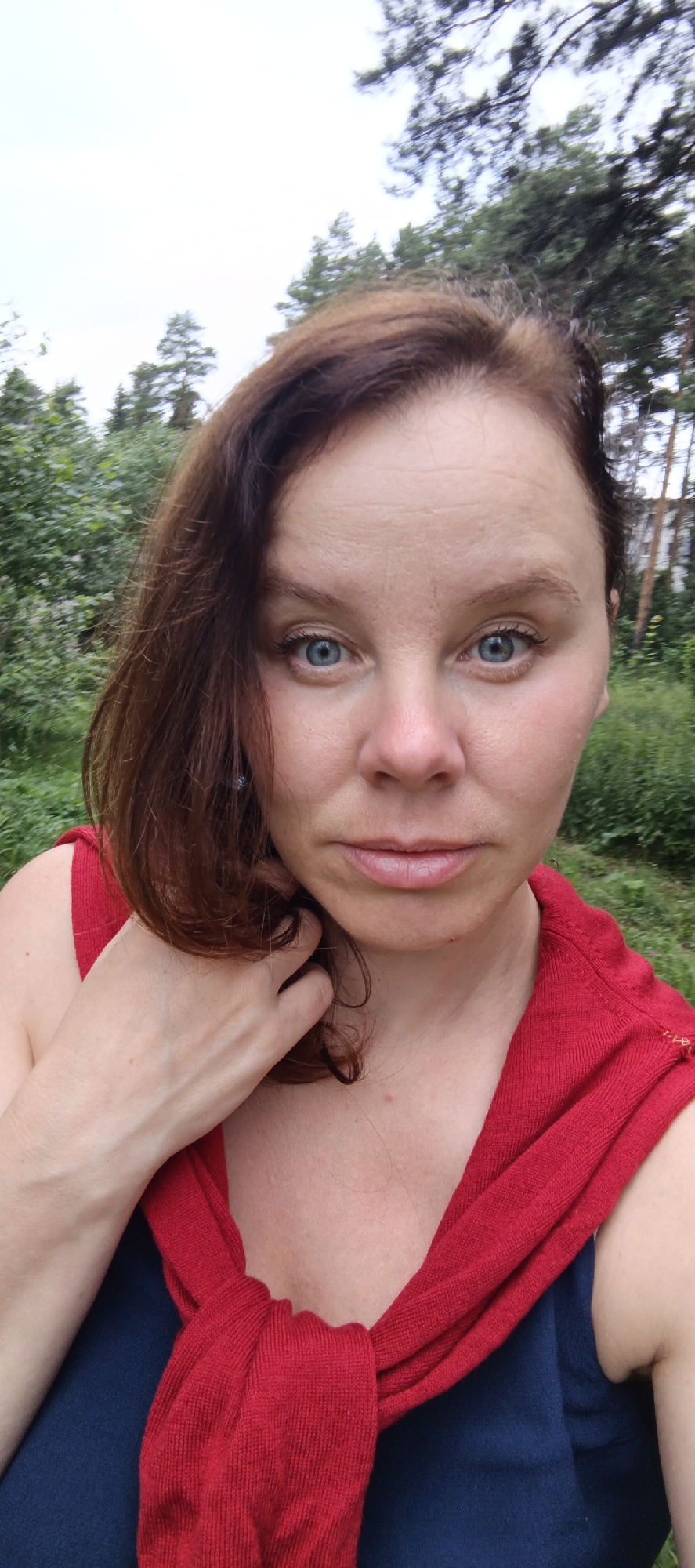 A woman with light skin, auburn hair, and striking blue eyes is looking directly at the camera. She is outdoors, with a blurred background of green foliage and tall trees under an overcast sky, suggesting a cloudy day. She is wearing a red garment draped around her shoulders over a dark blue top. Her left hand is raised, with fingers gently touching her hair near her face. Her expression is neutral, with a slight hint of a pensive or curious emotion. The setting appears to be a natural environment, possibly a park or forest, given the greenery and trees. The lighting is soft, typical of an overcast day, and there is no visible text in the image.
