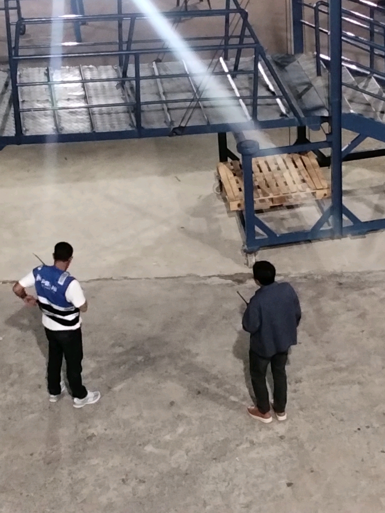 Two men are standing on a concrete floor in what appears to be an industrial or warehouse setting. One man, on the left, is wearing a white t-shirt, a blue safety vest with white stripes, black pants, and white athletic shoes. He is holding a walkie-talkie and facing away from the camera. The other man, on the right, is wearing a dark blue jacket, black pants, and brown shoes. He is also holding a walkie-talkie and facing away from the camera. In the background, there is a large blue metal structure with metal grating and wooden pallets. The lighting suggests an indoor environment, possibly with overhead lights creating bright glare. The scene is devoid of any specific markings or context that would definitively place it in Batam, Indonesia, or any other specific city.