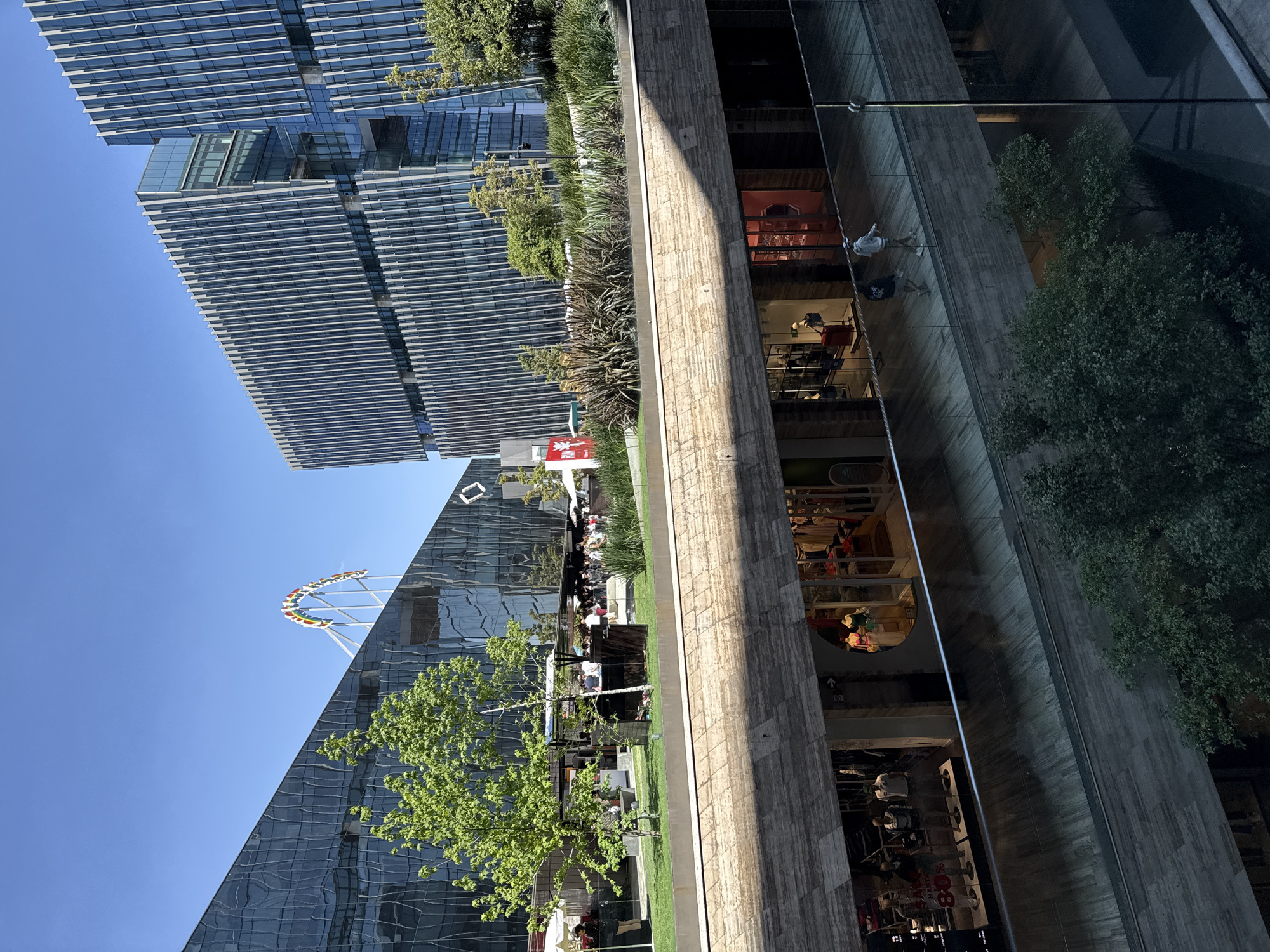 The image depicts an outdoor urban scene in Mexico City, Mexico. Two tall, modern buildings with glass facades dominate the background. One building features a Ferris wheel on its roof. Between the buildings is a green space with trees, grass, and people. There are some tables and chairs set up, suggesting a dining or gathering area. In the foreground, a multi-story building with a textured facade has storefronts visible through large glass windows, displaying commercial goods and people inside. A reflection of the adjacent building is partially visible on the large glass windows in the building on the right. The sky is clear and blue.