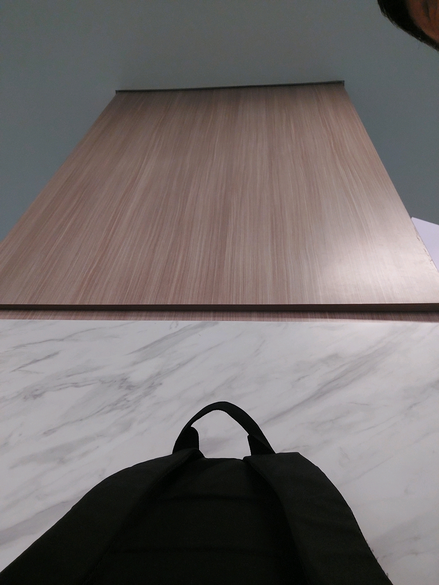 The image shows a close-up, low-angle perspective of a black backpack on a marble-patterned surface, with a light brown wooden panel above. The top edge of the wooden panel is visible against a pale blue wall. The scene appears to be indoors, possibly a room or an office space, in an unspecified location in Bekasi, Indonesia. There are no people or explicit actions depicted, only inanimate objects and a static environment. The lighting suggests an indoor setting with artificial or diffused natural light.