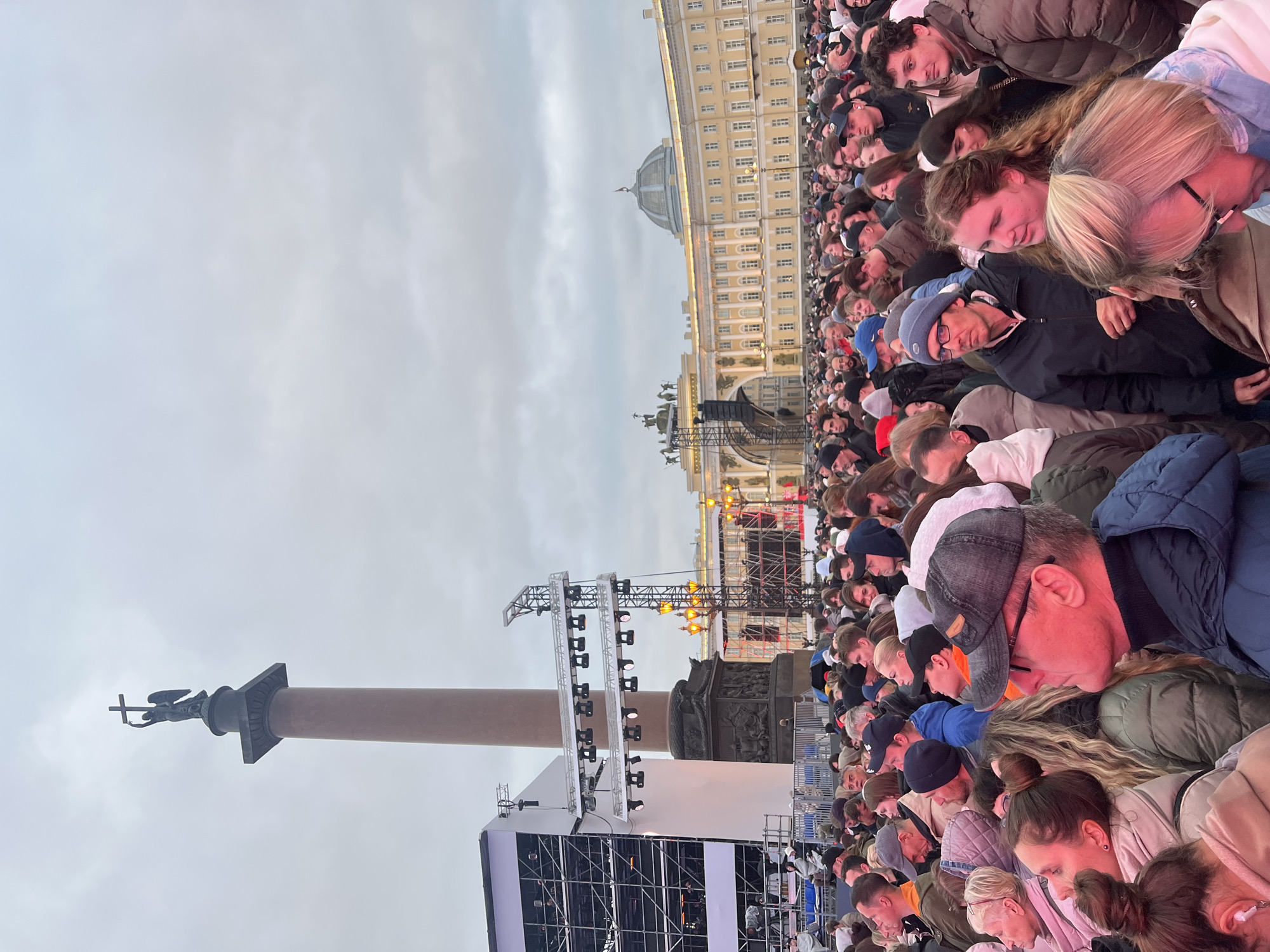 The image shows a large crowd of people gathered in Palace Square in Saint Petersburg, Russia. In the background, the Winter Palace and Alexander Column are visible. The crowd appears to be watching a stage set up in the square, likely for an event.