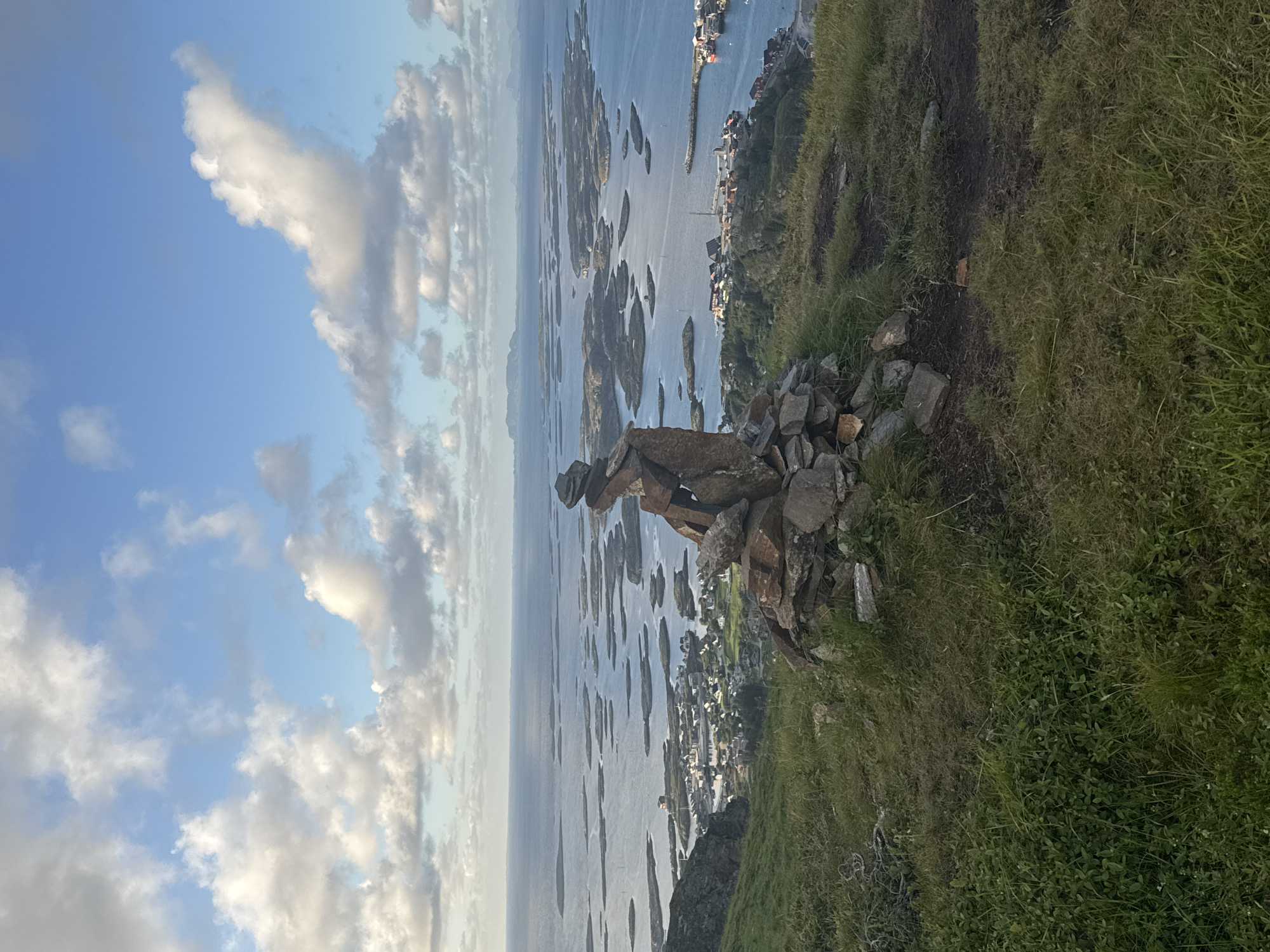 An outdoor, horizontal, eye-level, long shot captures a cairn of stacked stones in the foreground, positioned on a grassy hill overlooking a coastal town and a vast expanse of the ocean dotted with numerous small islands. The sky above is a bright blue, adorned with scattered, fluffy white clouds. The town, nestled along the shoreline, features a collection of buildings with red roofs and a harbor with several boats. The water is calm and reflects the light. The overall scene conveys a sense of natural beauty and tranquility. Based on the distinctive landscape and the presence of a prominent rock formation, this image is likely taken on the island of Lovund, Norway, a location known for its rugged coastline and abundant bird life.