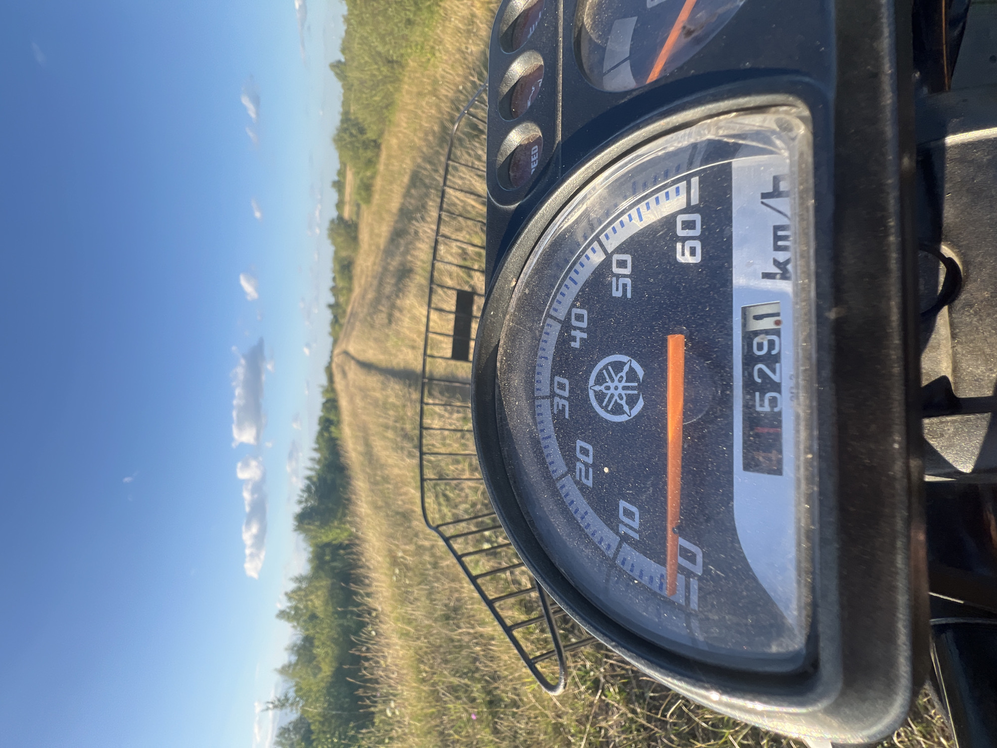 The image is a first-person perspective of someone riding a Yamaha scooter along a dirt road in the countryside near Sobishchytsi, Ukraine. The foreground is dominated by the scooter's dashboard, which includes a speedometer indicating a speed of approximately 45 km/h and an odometer reading of 4529.1 km. A black metal basket is mounted on the front of the scooter. The background shows a rural landscape with dry grass, scattered trees, and a clear blue sky with a few scattered clouds. The road appears to be unpaved and winds through the grassy fields. The lighting suggests it is daytime, likely late afternoon given the long shadows.