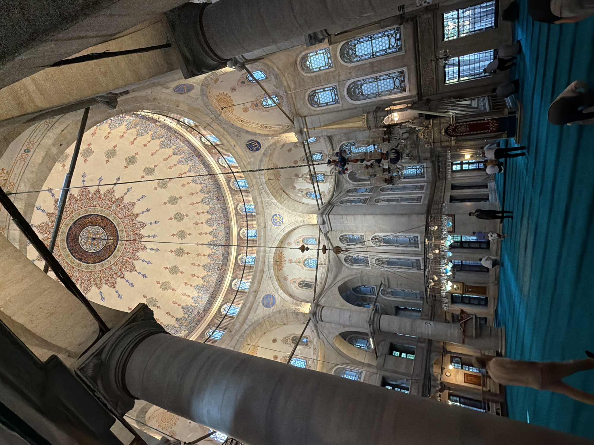 The image captures the interior of the Eyüp Sultan Mosque in Eyüpsultan, Turkey, a significant religious site. The viewpoint is from within the main prayer hall, looking upwards towards the grand dome and across the expanse of the floor.
Dominating the upper portion of the image is the mosque's impressive dome, adorned with intricate and colorful geometric patterns and calligraphy. The dome is supported by massive, classically styled columns, one of which is prominently visible in the left foreground, showing its fluted texture and substantial girth. The walls are lined with arched windows, many of which are tall and narrow, letting in natural light that creates a soft illumination within the space. Beneath the dome, arched alcoves and decorative motifs further enhance the architectural grandeur.
The floor of the prayer hall is covered with a vibrant, uniform teal carpet, indicative of a place of worship where congregants perform *salat* (prayer). Several individuals are present within the hall, engaged in various activities. Some are standing or walking, while others are in the process of prayer, some bowed in *ruku'* (bowing) and others in *sujud* (prostration). The figures are scattered across the carpeted area, suggesting a moderate presence of worshippers.
Large, ornate chandeliers hang from the ceiling, providing additional lighting. In the mid-ground, a prominent minbar (pulpit) is visible, a key feature in any mosque's prayer hall. The overall atmosphere appears serene and reverent, typical of a mosque during a period of prayer. The architecture speaks to a rich history and tradition, with elements of Ottoman design evident in the decorative schemes and structural forms.