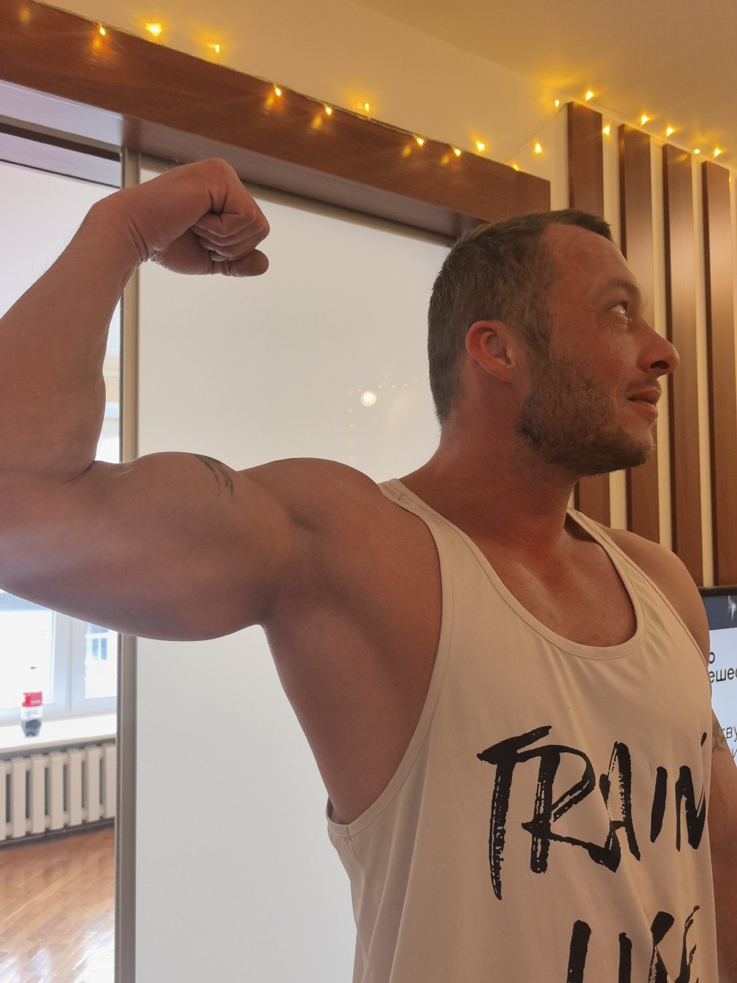 A man is posing indoors, flexing his left arm to display his bicep. He is wearing a white tank top with the words "TRAIN LIKE" printed on it in black. He is fair-skinned with short brown hair and a trimmed beard. He is looking to his right. The background includes a white sliding door, a window with a view of a radiator and a building exterior, and a wall decorated with vertical wooden slats and string lights. The location is Nizhny Novgorod, Russia, as evidenced by Cyrillic text visible on a screen behind the man.