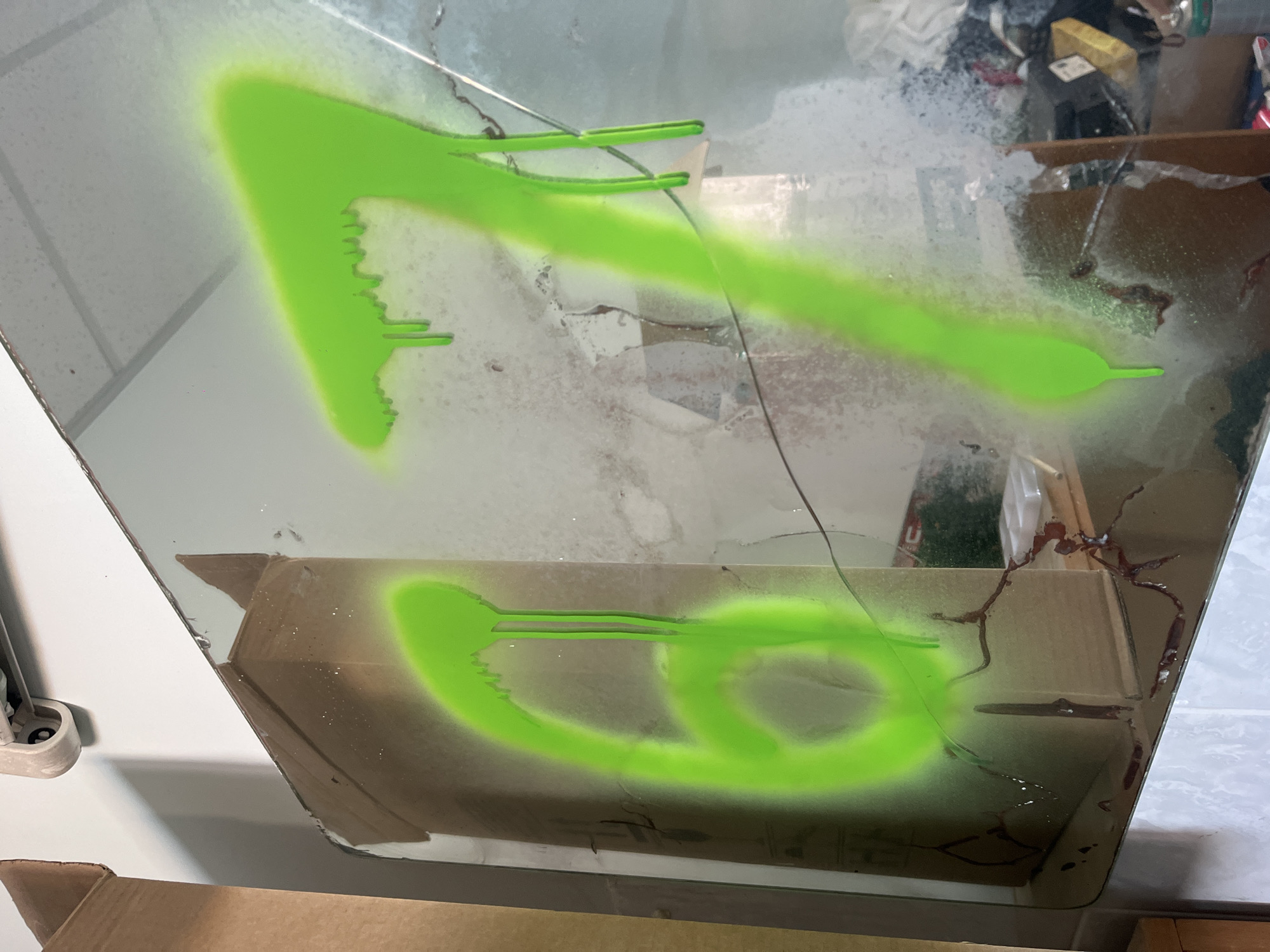 A damaged mirror is shown indoors. The mirror is cracked in several places and has been spray-painted with green paint in two distinct designs: an arrow shape near the top, and a rounded shape near the bottom. A cardboard box is partially visible behind the mirror. The room contains various objects including electrical equipment, various boxes and containers, and what appears to be a white wall. The location is not evident from the image and the claim that this is located in Nova Zagora, Bulgaria, is not substantiated by the image.