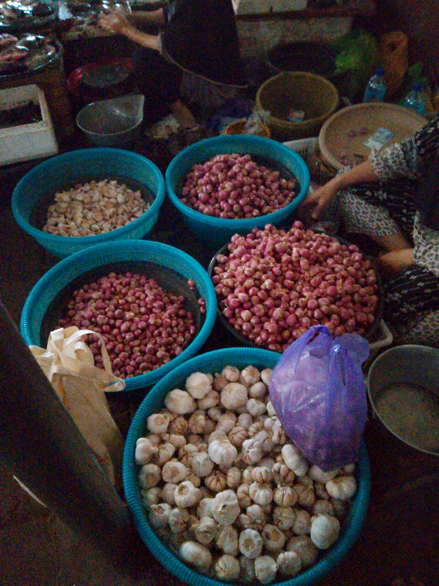 This is a high-angle, medium shot image taken at a market in Kediri, Indonesia. The scene depicts a display of fresh produce, primarily onions and garlic, arranged in blue plastic baskets on the ground. In the foreground, a large blue basket is filled with garlic bulbs, with a purple plastic bag partially covering some of them. To the left, a tan paper bag with a ribbon is also visible. In the mid-ground, a blue basket overflows with small red onions, and another basket to its left contains a lighter-colored vegetable, possibly shallots or dried garlic. Further back, another basket of red onions is seen. In the background, larger bowls or baskets are present, containing more produce, and a person's hand is visible reaching towards the produce, suggesting a vendor or shopper interacting with the items. The overall environment appears to be an outdoor or semi-outdoor market setting, with a dimly lit and somewhat cluttered atmosphere.