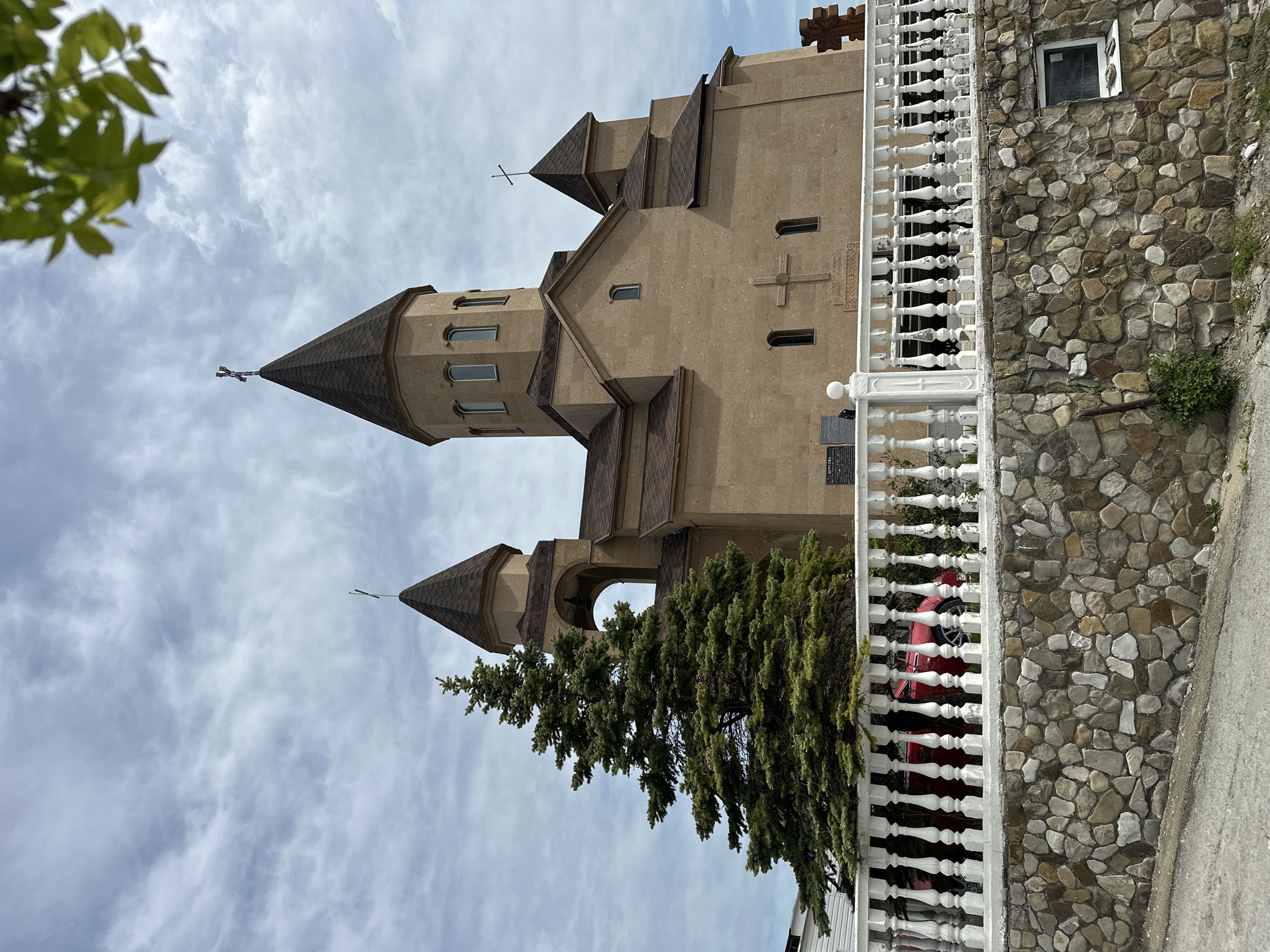 The image depicts a beige church with brown trim in Novomikhaylovsky, Russia. The church has two smaller towers and a larger tower with a spire topped with a cross. There are small crosses etched into the facade of the church. A white balustrade fence runs along the right side of the image. Behind the fence is a stone wall with a small, open window. A pine tree obscures part of the church. A red vehicle is partially visible behind the fence. The sky is cloudy. Green leaves are visible in the upper left corner. The image is vertically oriented.