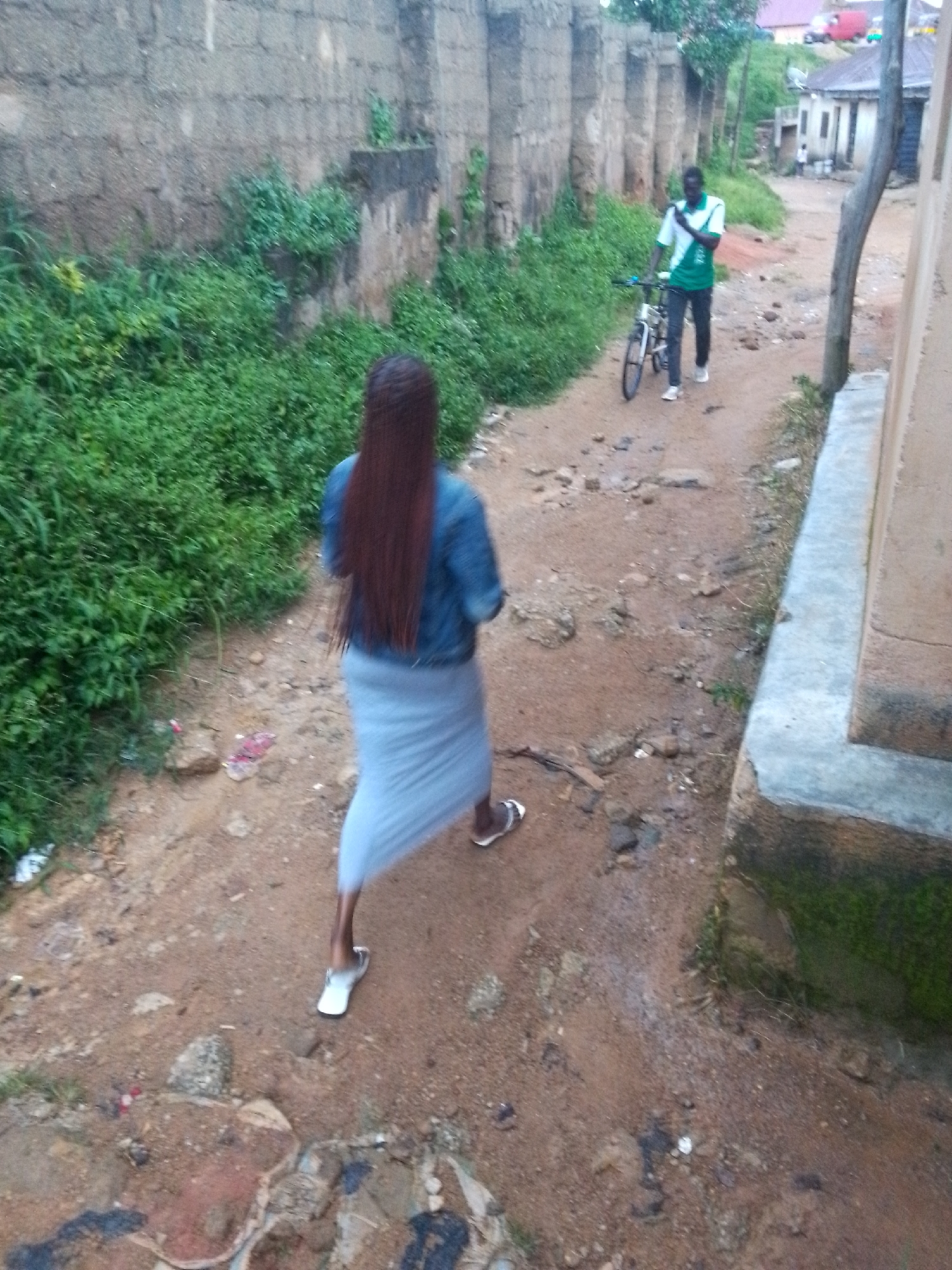 In Jos, Nigeria, a young woman with long, braided, reddish-brown hair walks away from the camera, wearing a blue denim jacket and a light blue, flowing skirt. She has white flip-flops on her feet. Beside her, a man in a green and white t-shirt and dark pants walks a bicycle. The scene unfolds on a dirt path lined with lush green vegetation and a rough, grey stone wall. In the background, a few buildings are visible, suggesting a residential area. The overall environment appears somewhat unkempt, with debris scattered on the path.