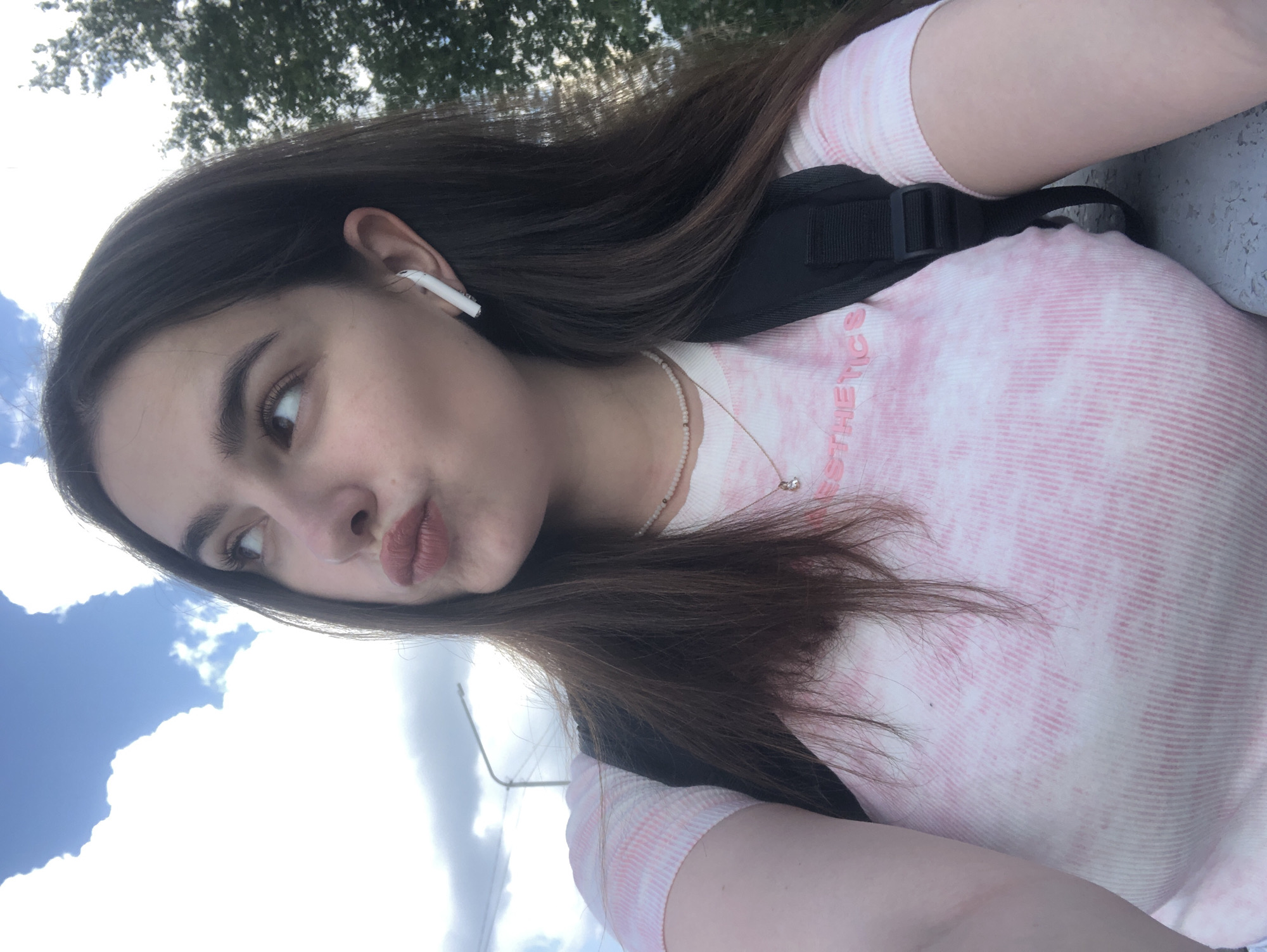 A young woman with long, dark brown hair is taking a selfie outdoors. She is wearing a pink and white tie-dye t-shirt and a black backpack. She has on a delicate necklace with small beads and a pendant. There is a white wireless earbud in her right ear. Her lips are pursed as she looks off to the side. The background shows a cloudy blue sky and the tops of green trees, suggesting an urban park or street setting. A streetlight is visible in the background to the left. Based on the distinctive style of the streetlight, this location is likely Chaikovsky, Russia.