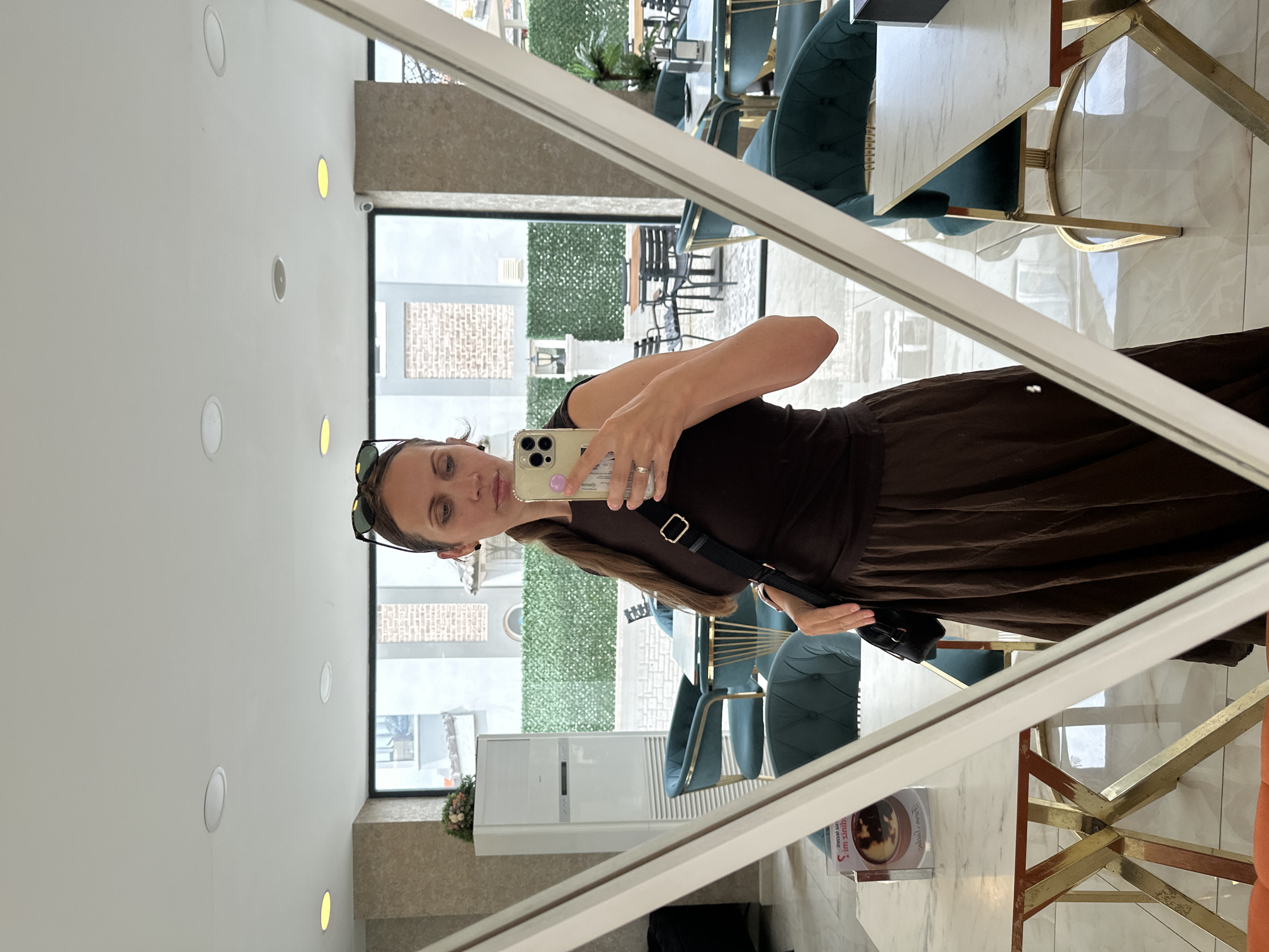 The image is a mirror selfie, captured by a woman using a smartphone, and is presented rotated 90 degrees counter-clockwise. The woman, who has brown hair and is wearing a dark brown top and what appears to be a matching skirt, is looking at her phone. Sunglasses are pushed up onto her head, and a black shoulder bag strap crosses her body. Her left hand holds a light-colored smartphone (possibly gold or beige), which is being used to take the picture. A silver ring is visible on her left ring finger.
The selfie reflects a modern cafe or restaurant interior. The visible furniture includes teal, upholstered dining chairs with gold-colored metal frames and tables featuring white marble tops also supported by gold-colored metal bases. The floor appears to be light-colored tile. Recessed circular lights, some emitting a warm yellow glow and others unlit, are visible on the white ceiling.
Through large windows in the background, an outdoor area is visible. This area features green artificial hedges, dark outdoor seating (chairs and tables), and what appears to be a paved or tiled ground. Beyond the immediate outdoor space, other buildings with varying architectural styles can be seen. A white, modern air conditioning unit is positioned near one of the windows, and a security camera is mounted above another window frame. A small decorative potted plant is also visible near a window. On one of the marble tables, a white item, possibly a menu or flyer, is present, displaying the partial text "Ups!" and "Turkish" or "Turk...". The setting appears to be bright and well-lit, indicative of daytime. Given the "Turkish" text, the location context of Fethiye, Turkey, is reinforced.