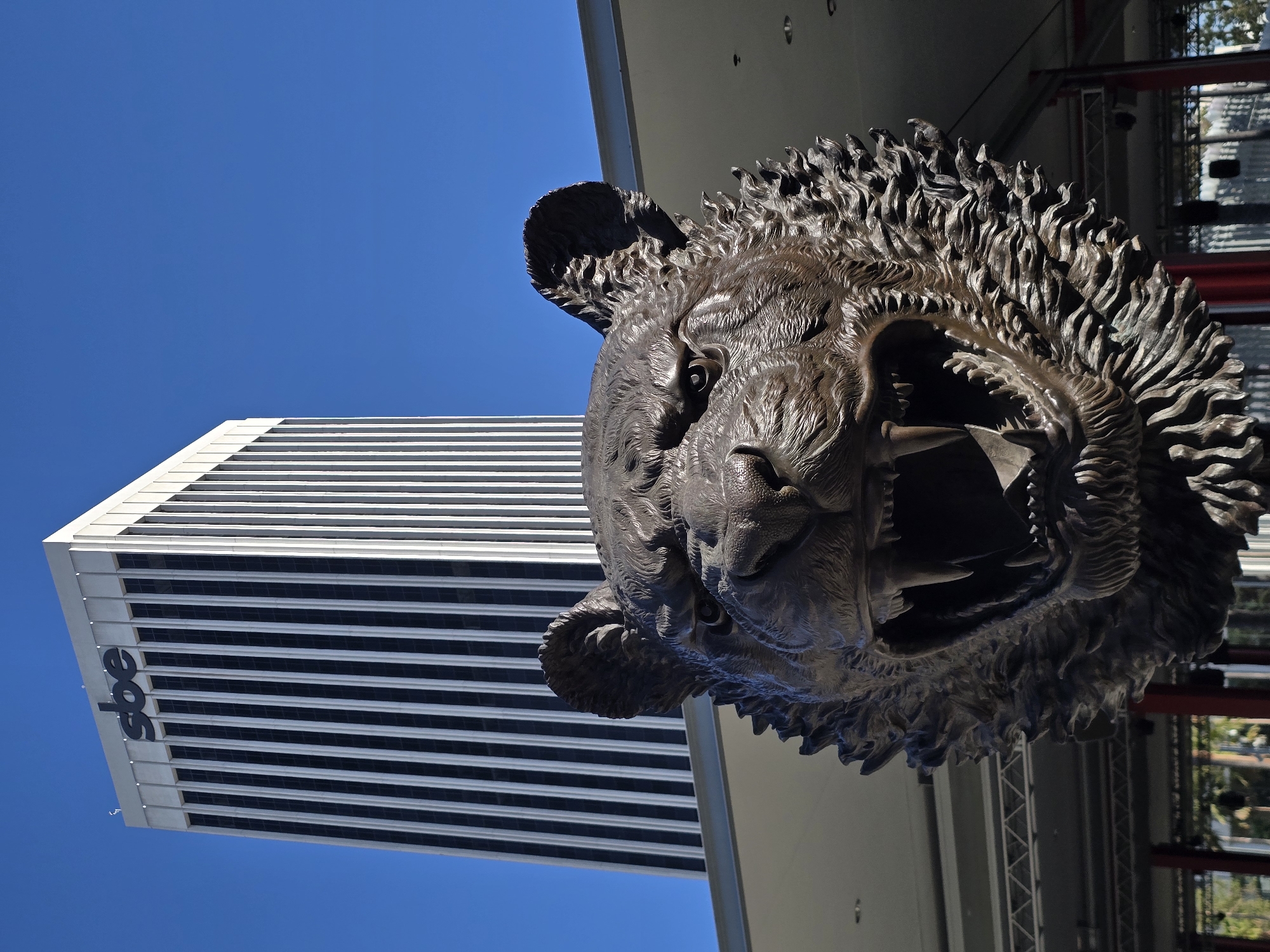 A bronze sculpture of a snarling tiger's head is positioned in the foreground, facing left, with its mouth open revealing sharp teeth. Behind the sculpture, a modern, white skyscraper with a dark, vertical-striped facade rises against a clear blue sky. The SBE logo is visible at the top of the building. The sculpture is mounted on a horizontal structure, possibly a balcony or overhang, with a light-colored ceiling visible above it. In the background, beneath the overhang, some scaffolding and red poles suggest an outdoor event space or construction. The scene appears to be in a city environment, with glimpses of greenery in the far distance.