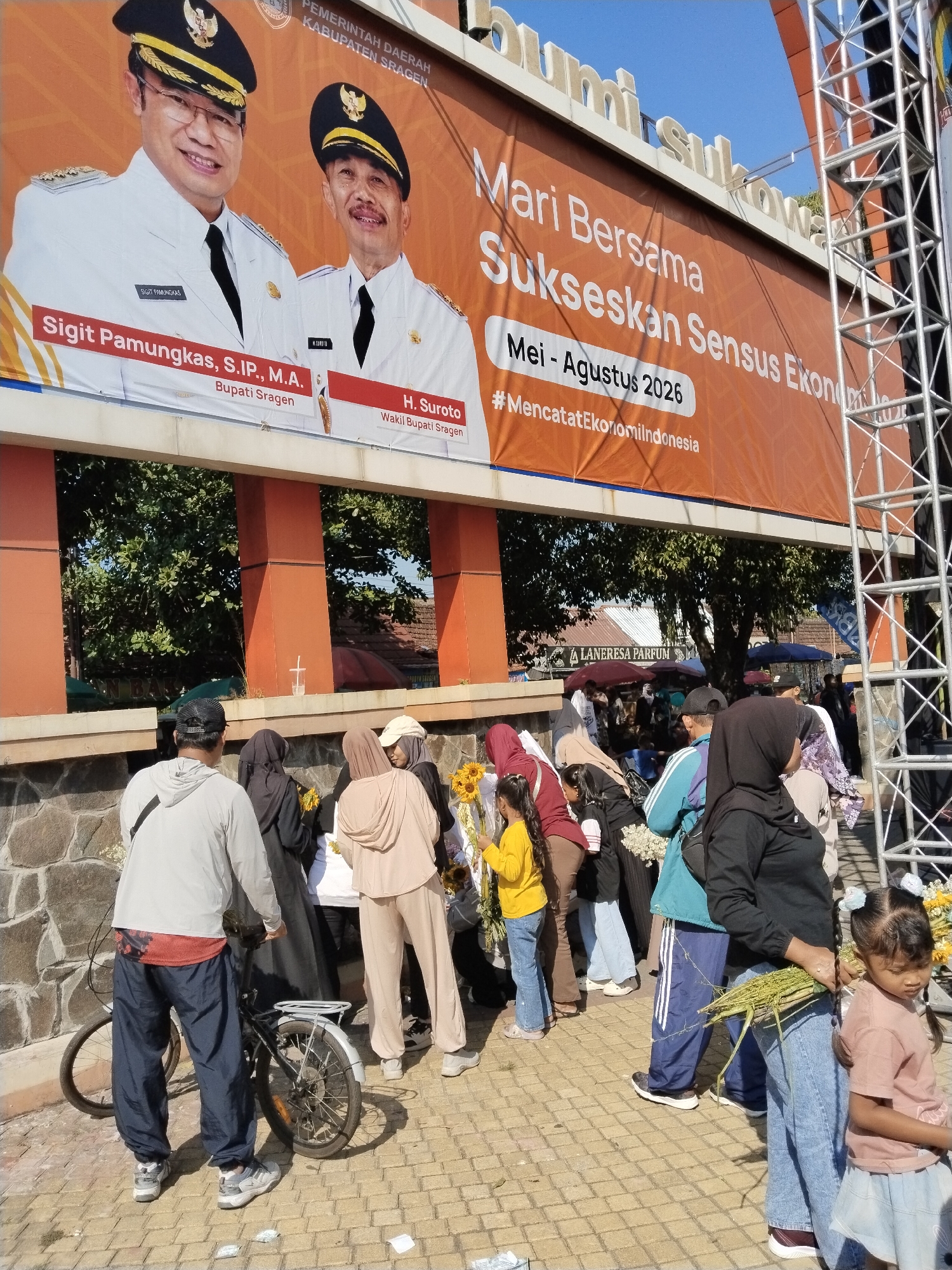 The image depicts an outdoor public scene in Sragen Regency, Indonesia, characterized by a large banner promoting the 2026 Economic Census and a gathering of people.
At the top, a prominent orange and white banner displays "PEMERINTAH DAERAH KABUPATEN SRAGEN" (Local Government of Sragen Regency). It features two smiling men in white civil servant uniforms and black caps: Sigit Pamungkas, S.I.P., M.A., identified as the Regent of Sragen, and H. Suroto, identified as the Vice Regent of Sragen. The banner's main text reads "Mari Bersama Sukseskan Sensus Ekonomi" (Let's Together Succeed the Economic Census) with the timeline "Mei - Agustus 2026" (May - August 2026) and the hashtag "#MencatatEkonomiIndonesia" (Recording Indonesia's Economy). Above the banner, white 3D letters spelling "Bumi Suka..." are visible, part of "Bumi Sukowati," a nickname for Sragen.
Below the banner is an orange-brown architectural structure with rectangular openings revealing green foliage and a bright blue sky. To the right, a metal scaffolding structure ascends vertically.
In the mid-ground and foreground, numerous individuals are gathered on a patterned yellow/tan paved ground. The majority are adults, with several women wearing headscarves (hijabs) and modest clothing. Some people are holding items such as bouquets of sunflowers and bundles of long green leaves.
On the far left, a man seen from behind wears a light grey hooded sweatshirt, dark blue track pants with a red pattern visible at the waist, and grey sneakers. He stands next to a black bicycle equipped with a rear rack and fenders. In the center, a woman in a beige hooded outfit holds sunflowers, while another woman in a black hijab holds green leaves. A young girl with pigtails, wearing a pink shirt and blue skirt, stands to the right, looking downwards. Another girl in a yellow shirt and blue jeans, holding sunflowers, is partially visible further left.
In the background, under an awning, a sign reading "LANERESA PARFUM" is visible, suggesting a commercial area or market stall. Scattered small white pieces of paper or plastic are on the paved ground in the immediate foreground.