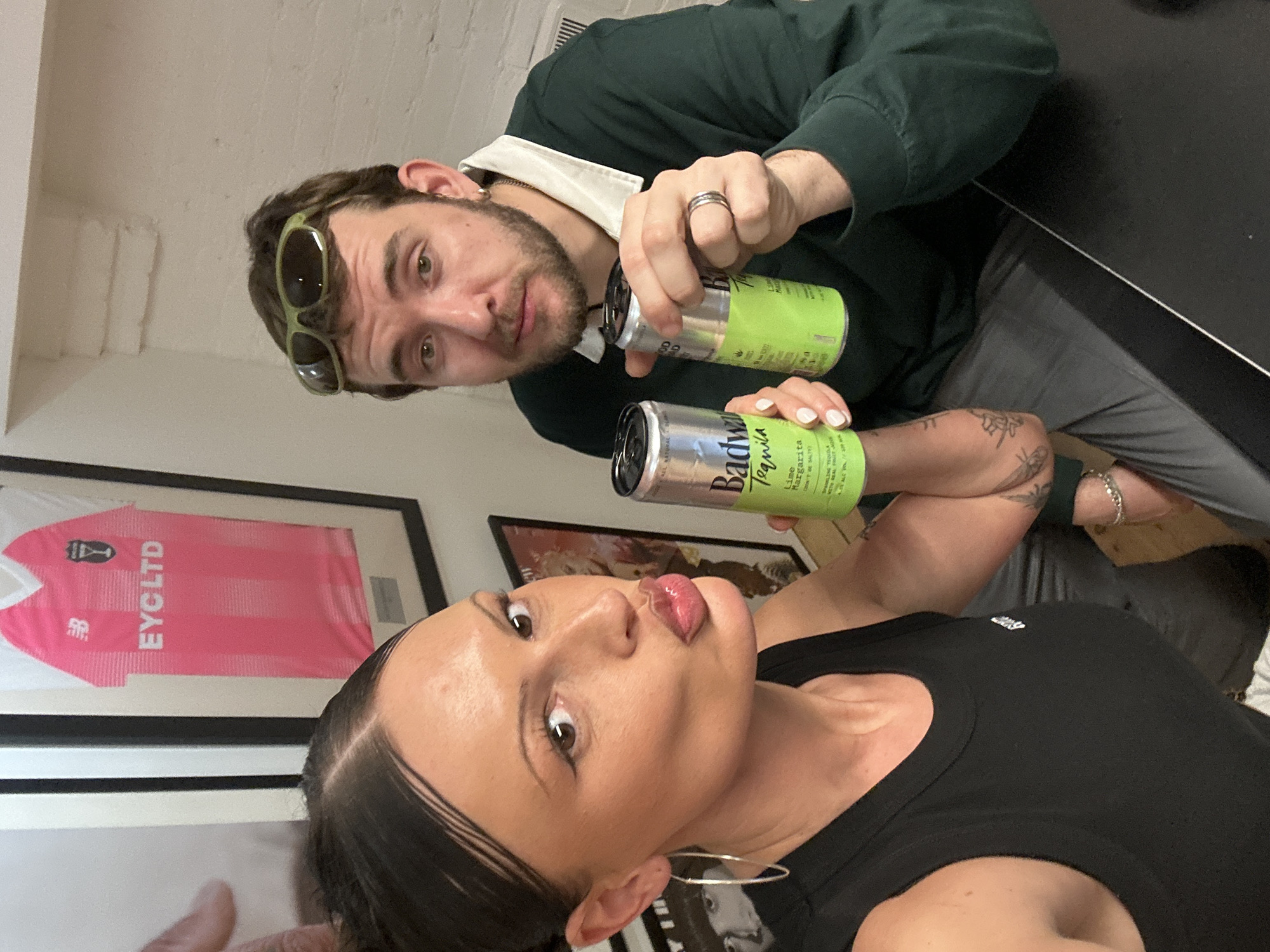 The image depicts two individuals, one female and one male, indoors, posing for a photograph. The scene is situated in London, United Kingdom.
The female, positioned on the left, has dark hair styled back and is wearing a black tank top and large silver hoop earrings. She has white nail polish on her visible fingernails and multiple tattoos on her left forearm/bicep area. She is looking directly at the camera with her lips pursed in a pouting expression, holding a silver and lime-green can of "Badwahl Tequila Lime Margarita" in her left hand, which specifies "Lime Margarita" and "5% ALC VOL".
The male, positioned on the right, has light brown hair, a beard, and green sunglasses resting on top of his head. He is wearing a dark green long-sleeved polo shirt with a white collar, a silver earring in his left earlobe, and a silver ring on his left ring finger. He is also looking directly at the camera with a neutral expression, holding an identical "Badwahl Tequila Lime Margarita" can in his left hand.
The background features a white painted wall. A framed pink football jersey is prominently displayed, showing a New Balance logo, the text "EYCLTD," and a crest resembling that of Liverpool FC. Below this, a portion of another framed item, possibly a black-and-white portrait or album cover, is visible. Architectural details suggesting brickwork and a radiator are partially discernible in the upper right background. The foreground shows a dark, flat surface, consistent with a table.