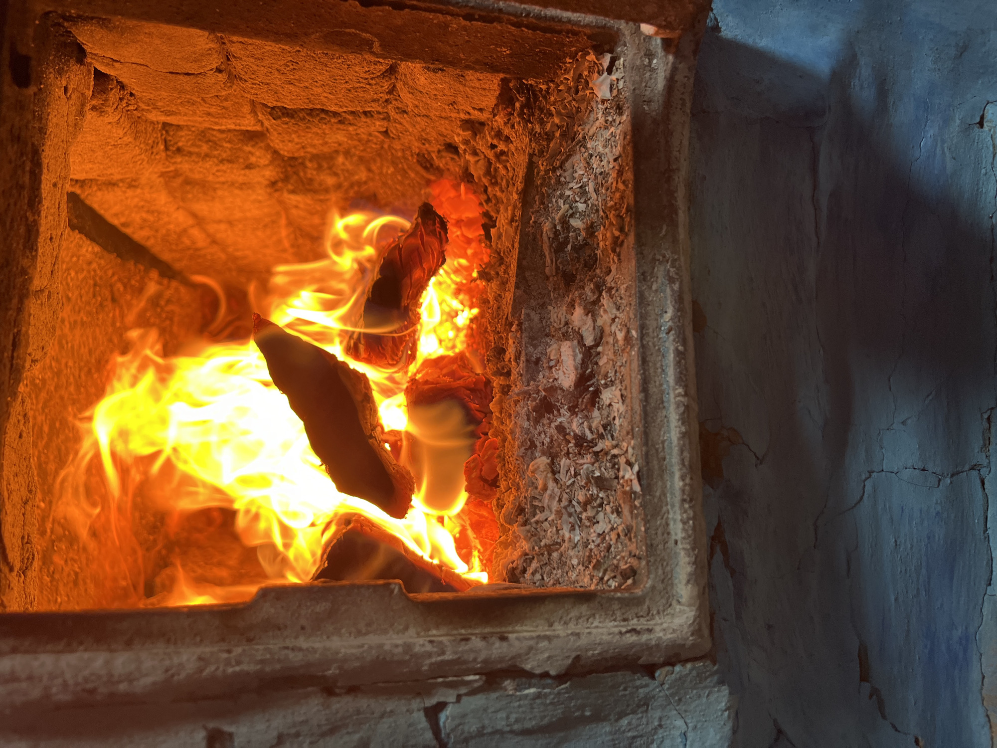 This image, taken in Nizhny Tagil, Russia, displays a detailed view into the active firebox of a wood-burning stove or fireplace. The central focus is a vibrant, multi-hued fire, dominated by intense yellow and orange flames that actively consume several wood logs positioned within the chamber. Below the main burning logs, a glowing bed of red and orange embers is visible, partially obscured by a substantial accumulation of light grey and whitish ash. The interior walls of the firebox, constructed from a rough-textured, possibly refractory, material, exhibit a strong orange-red coloration due to the intense heat and light emanating from the fire. These inner surfaces show evidence of wear and soot accumulation. The opening of the firebox is framed by a robust, dark-grey metallic or stone structure on the left and bottom, suggesting a sturdy, functional design. Below the firebox opening, the lower portion of the scene reveals a cracked, dull blue-grey wall or floor surface, indicating the immediate environment surrounding the heating appliance. No individuals or animate subjects are present in the frame. The primary action observed is the combustion of wood, producing light and heat.