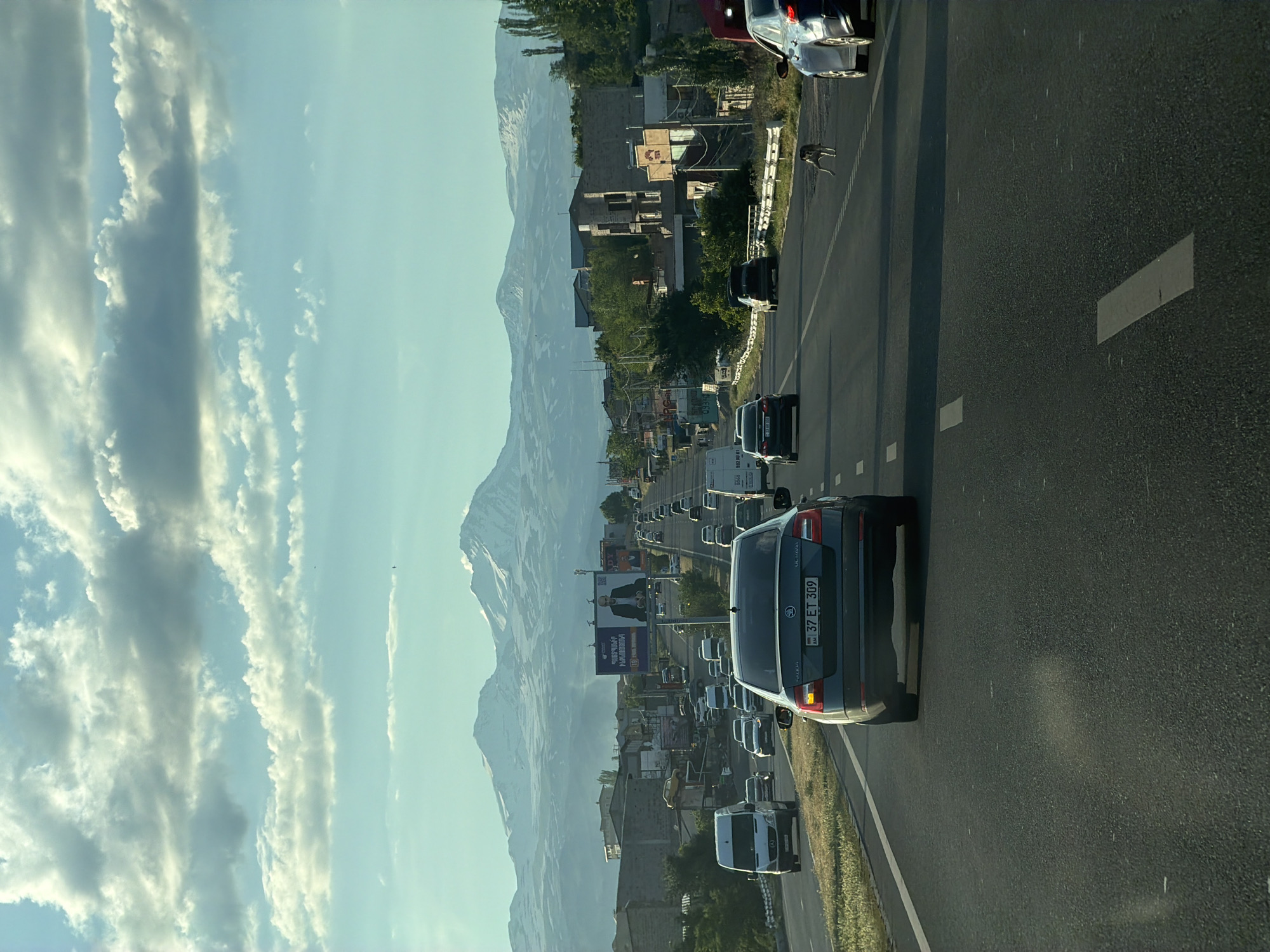 The image depicts a highway leading towards snow-capped mountains, with a scattering of buildings and billboards lining the sides of the road. The sky is filled with dramatic, cloudy formations. Several vehicles are visible on the highway, including a dark gray Skoda Octavia in the foreground with the license plate "37 ET 309". A lean dog is seen running across the highway in the middle distance. The environment suggests a semi-urban or suburban setting in Armenia, likely on the road leading to or from Proshyan, given the prominent billboards that may advertise local services or events. The lighting indicates it is either early morning or late afternoon.
