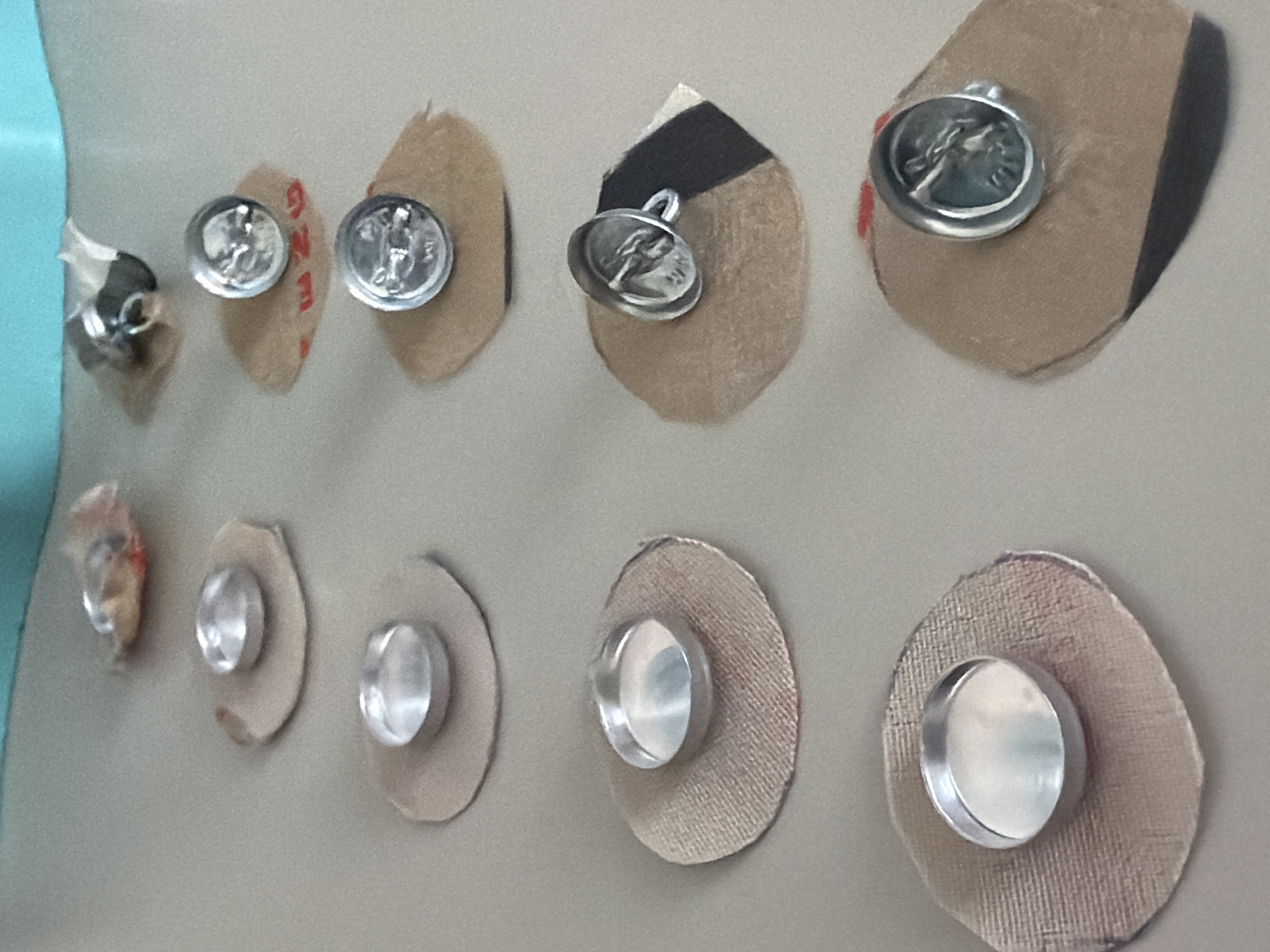 The image displays two rows of metallic objects affixed to light-colored, mostly beige or tan patches, arranged on a plain, light-colored background, likely a wall or display board. A light blue surface is visible on the far left edge.
In the upper row, there are approximately six patches, each bearing a silver-colored, coin-like object. These objects are detailed, featuring embossed classical profiles or figures, resembling ancient medallions or charms. One specific object has a small loop, suggesting it could be a pendant. Some of the patches in this row have additional markings; one clearly shows red text reading "GZM" oriented vertically, and another has a dark brown or black segment.
The lower row consists of about six simpler, smooth, silver-colored metallic discs or caps. These are attached to more consistently circular or oval-shaped textured beige patches.
The objects are arranged in a linear fashion, implying a collection, an art display, or a series of sensors or electrodes, possibly in a medical or experimental context given the "GZM" text and the appearance of some elements. The setting is indoors, with no discernible time of day or weather. This scene is located in Saint Petersburg, Russia.