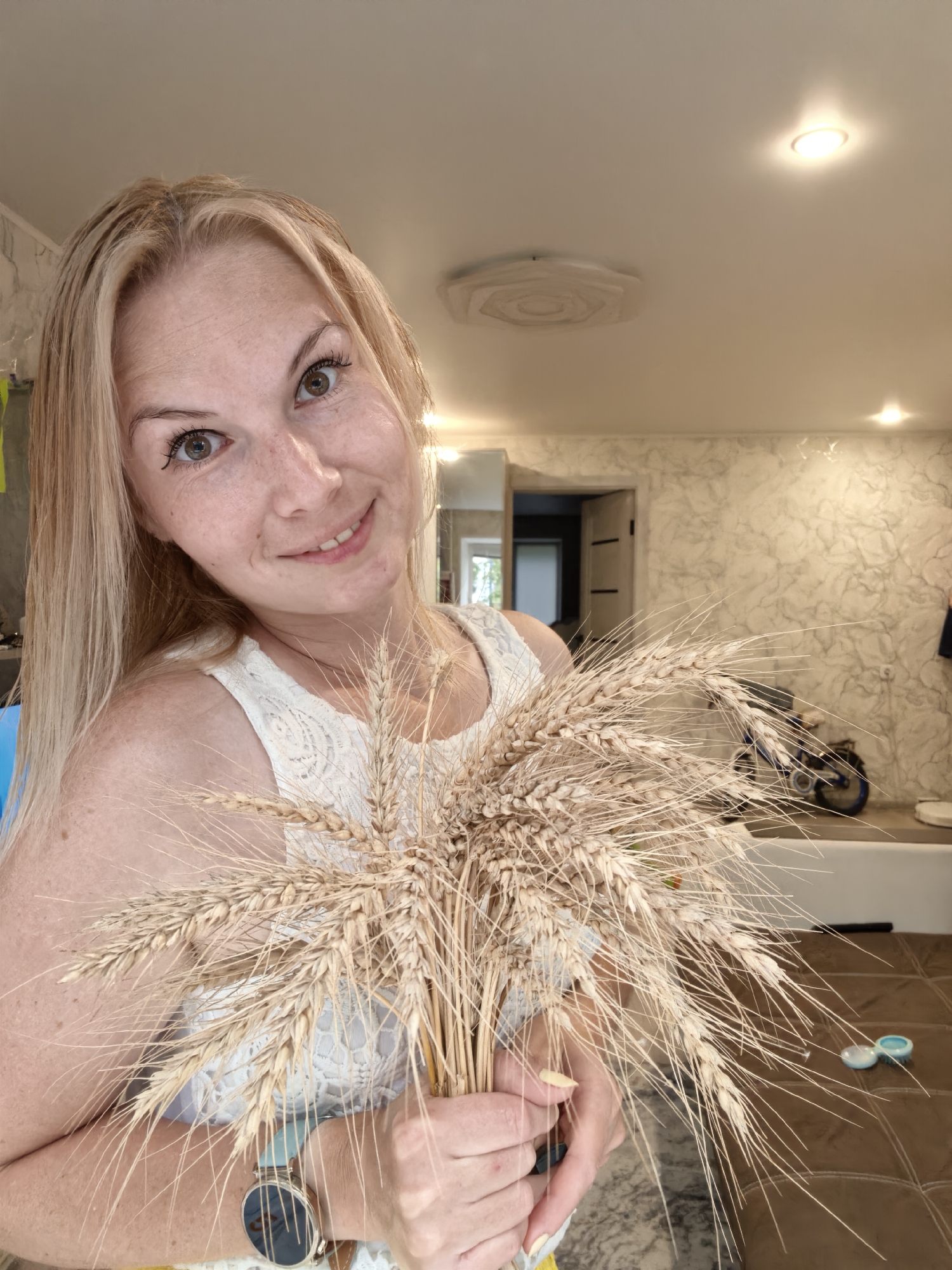 A blonde woman with brown eyes and freckles smiles at the camera, holding a bouquet of wheat stalks. She is wearing a white lace top and a watch on her left wrist. The background appears to be an indoor setting, possibly a home, with textured walls and a ceiling light fixture. In the background, a doorway leads to another room, and a bicycle is visible. The woman is positioned in the foreground, slightly to the left of the frame.