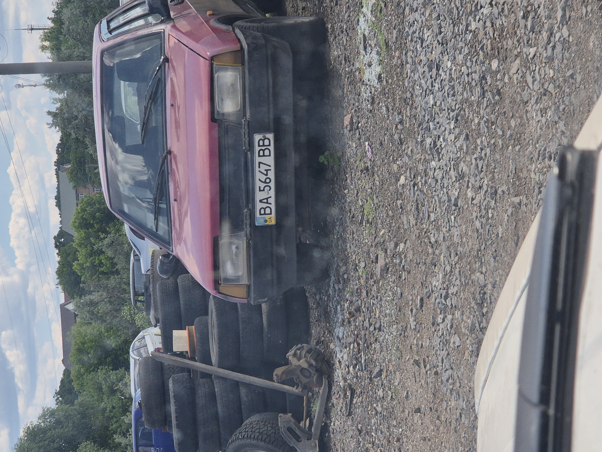 In Pidhaitsi, Ukraine, a red hatchback car with license plate BA 5647 BB is parked on a gravel lot, surrounded by stacks of used tires and car parts. The car's front bumper is dark and worn, with a yellowed headlight and a visible license plate holder. Behind the red car, other vehicles are partially visible, including a blue car and a white car. A few trees and a building with a reddish roof are seen in the background, under a cloudy sky. The scene appears to be an informal vehicle service area or junkyard.