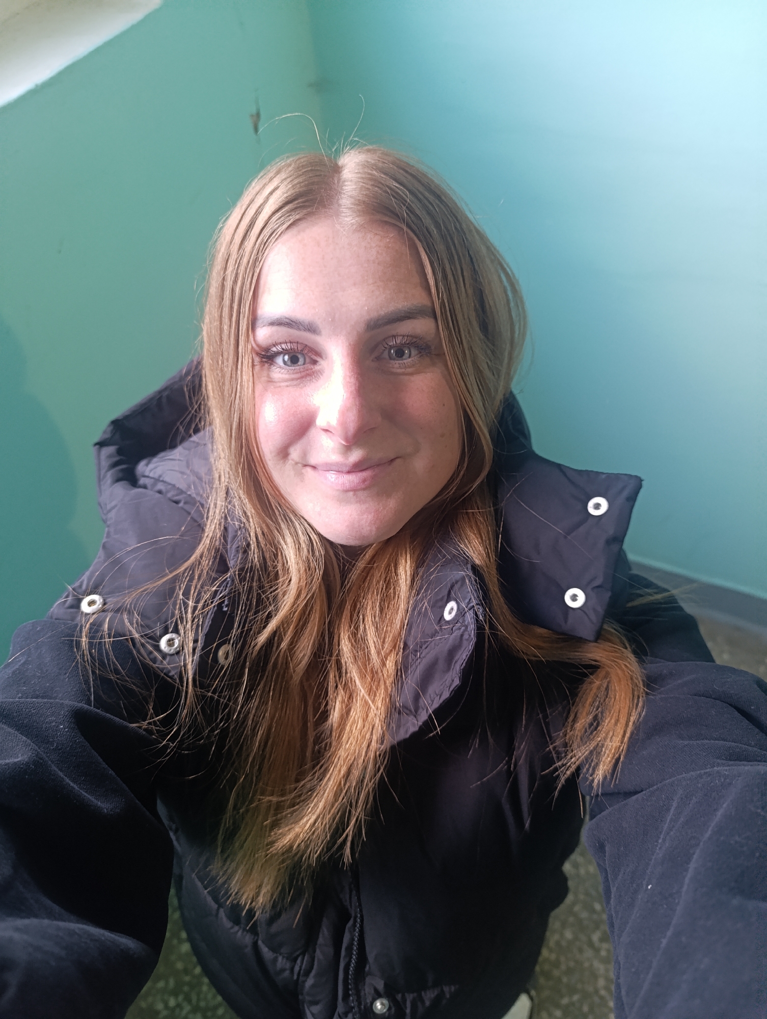 A selfie of a light-skinned young woman with light brown hair, freckles, and light-colored eyes, wearing a dark-colored, puffy winter coat with white snaps, against a light green wall in a city of Unknown, Unknown.