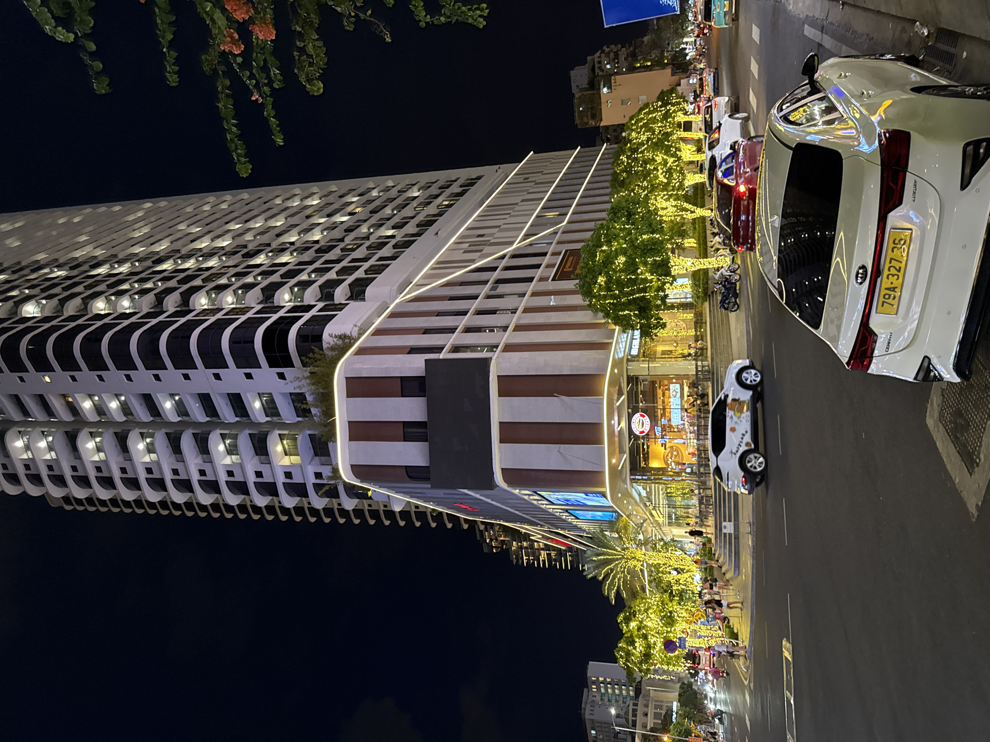 The image shows a city street at night in Nha Trang, Vietnam, with a prominent modern skyscraper illuminated by numerous lights. The skyscraper has a distinctive architectural design with curved balconies. In front of the skyscraper, there is a multi-story building with a facade featuring vertical stripes of white and a reddish-brown color, and it is decorated with bright yellow string lights wrapped around trees and along the building's edges. Several cars, including a white Kia Cerato with a yellow license plate "79A-327.35", are visible on the street. Pedestrians are seen walking on the sidewalk and near the entrance of the building, where a restaurant or cafe appears to be located. The overall atmosphere is vibrant and commercial, indicative of a busy urban area at night.