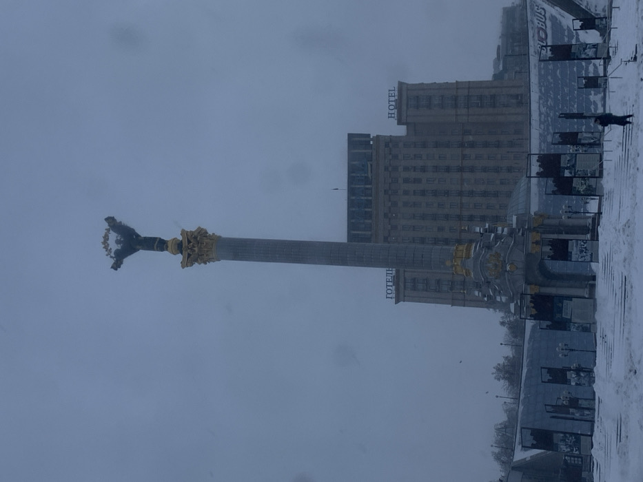 The image captures Maidan Nezalezhnosti (Independence Square) in Kyiv, Ukraine, under winter conditions. A prominent Independence Monument stands centrally, featuring a tall, fluted column with gold-colored decorative elements at its base and capital. A dark-colored statue of a female figure (Berehynia) holding a wreath is positioned atop the column.
Behind the monument, the multi-story Hotel Ukraina is visible, with its light-colored facade partially obscured by the ambient light. The words "ГОТЕЛЬ" (HOTEL) and "HOTEL" are affixed to the building's upper sections.
The ground and various architectural structures in the foreground are covered with a layer of undisturbed white snow. A series of snow-covered steps leads upwards toward the monument's base. Arranged in an arc on either side of the monument's lower section are several rectangular display structures, some appearing to feature photographic or textual content. A partial sign displaying "GLOBUS" is visible on the right, indicating an entrance to an underground complex.
A lone individual, dressed in dark attire, walks on the snow-covered steps in the lower right quadrant of the image. The sky is a uniform, overcast grey, indicative of a snowy or pre-snowfall atmosphere. Numerous small, bright specks, consistent with snowflakes or moisture on the camera lens, are present throughout the frame.