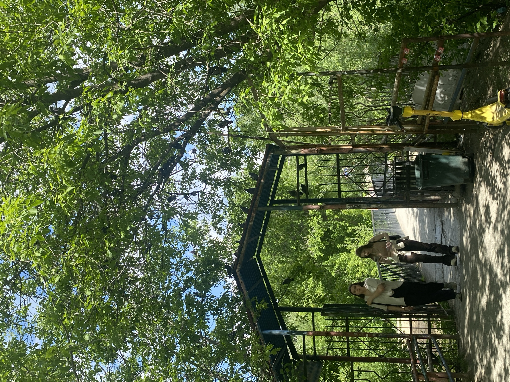 The scene depicts an outdoor area in Yekaterinburg, Russia. Two young women stand within a gated structure. One wears a white shirt and black pants, while the other is dressed in a tan top and dark brown pants. Green foliage is visible overhead, partially obscuring the sky. A yellow mechanical device is present near a trash can.