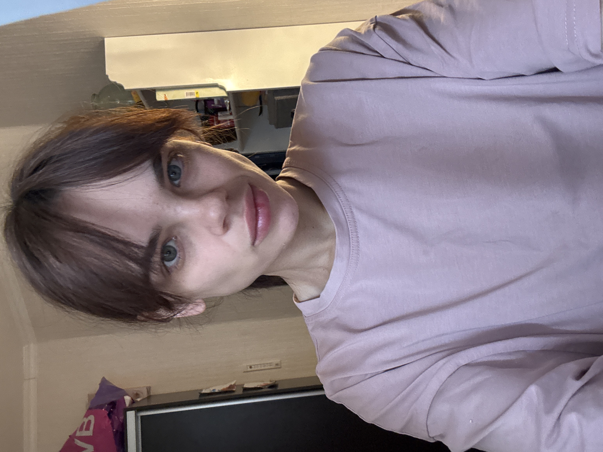 A woman with short, dark hair and blue eyes is centered in the frame, wearing a mauve-colored long-sleeved shirt. Her face is slightly angled towards the camera. The background is a blurred interior setting with light walls and a cluttered shelf. A dark object, possibly a television, is visible in the lower part of the image. No information suggests it is taken in Rostov-on-Don, Russia.