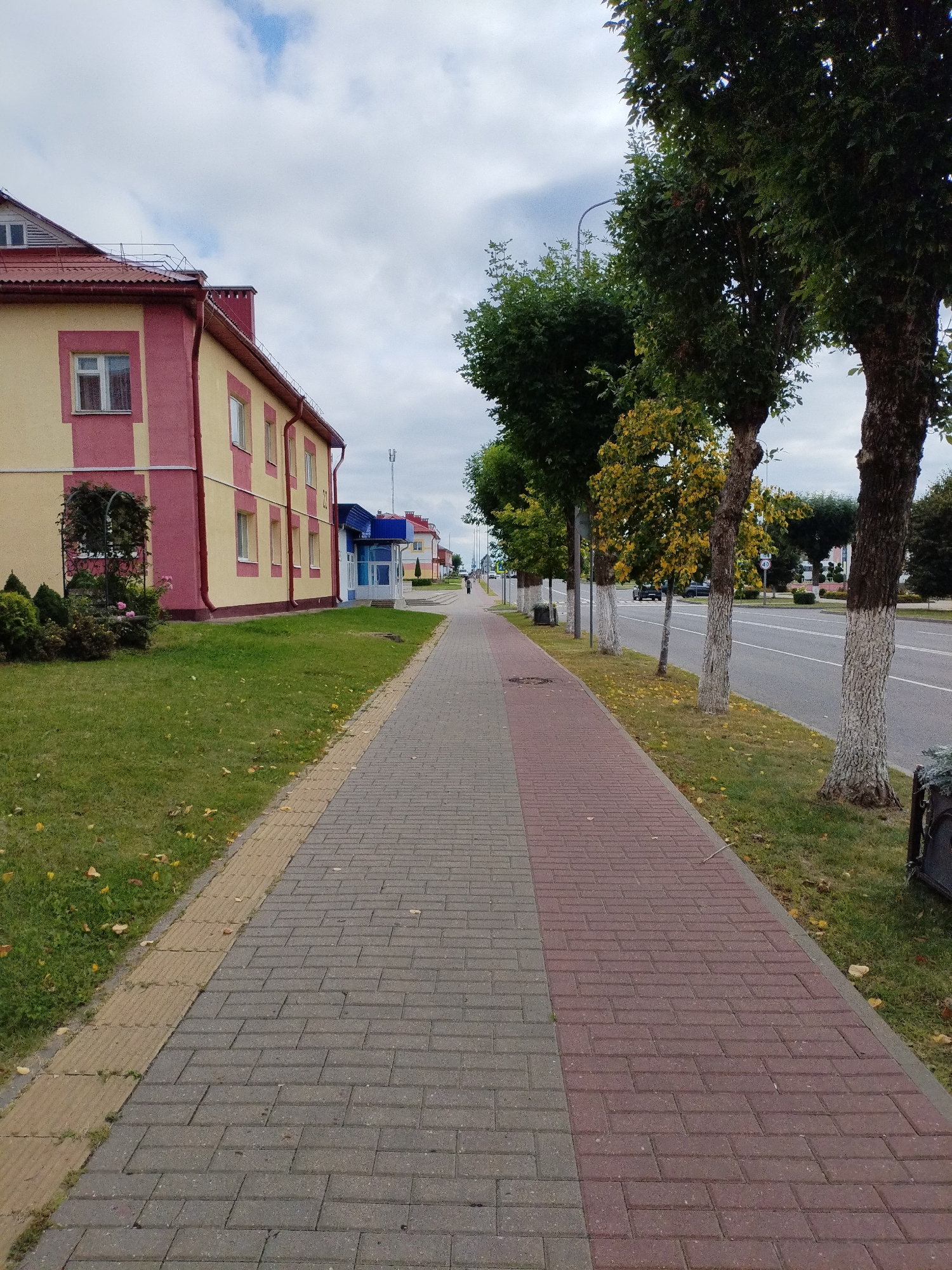 This is a medium shot, eye-level, outdoor, vertical, full-color photograph of a sidewalk running alongside a street in the city of Bialynichy, Belarus. The sidewalk is made of two alternating patterns of brick pavers, with a yellow tactile paving strip along the grassy edge. The street runs parallel to the sidewalk, with a row of trees lining the edge between them. The trees have white-painted trunks. On the left side of the sidewalk is a building with a light yellow facade and dark pink trim. It has several windows, and the roof is tiled in red. Further down the street on the left, other buildings are visible, including one with a blue roof and entrance. On the right side of the street, cars are visible driving and parked. The sky is overcast with scattered clouds. A lone pedestrian is visible walking in the distance on the sidewalk. The scene appears to be set in a town or city environment during daytime.