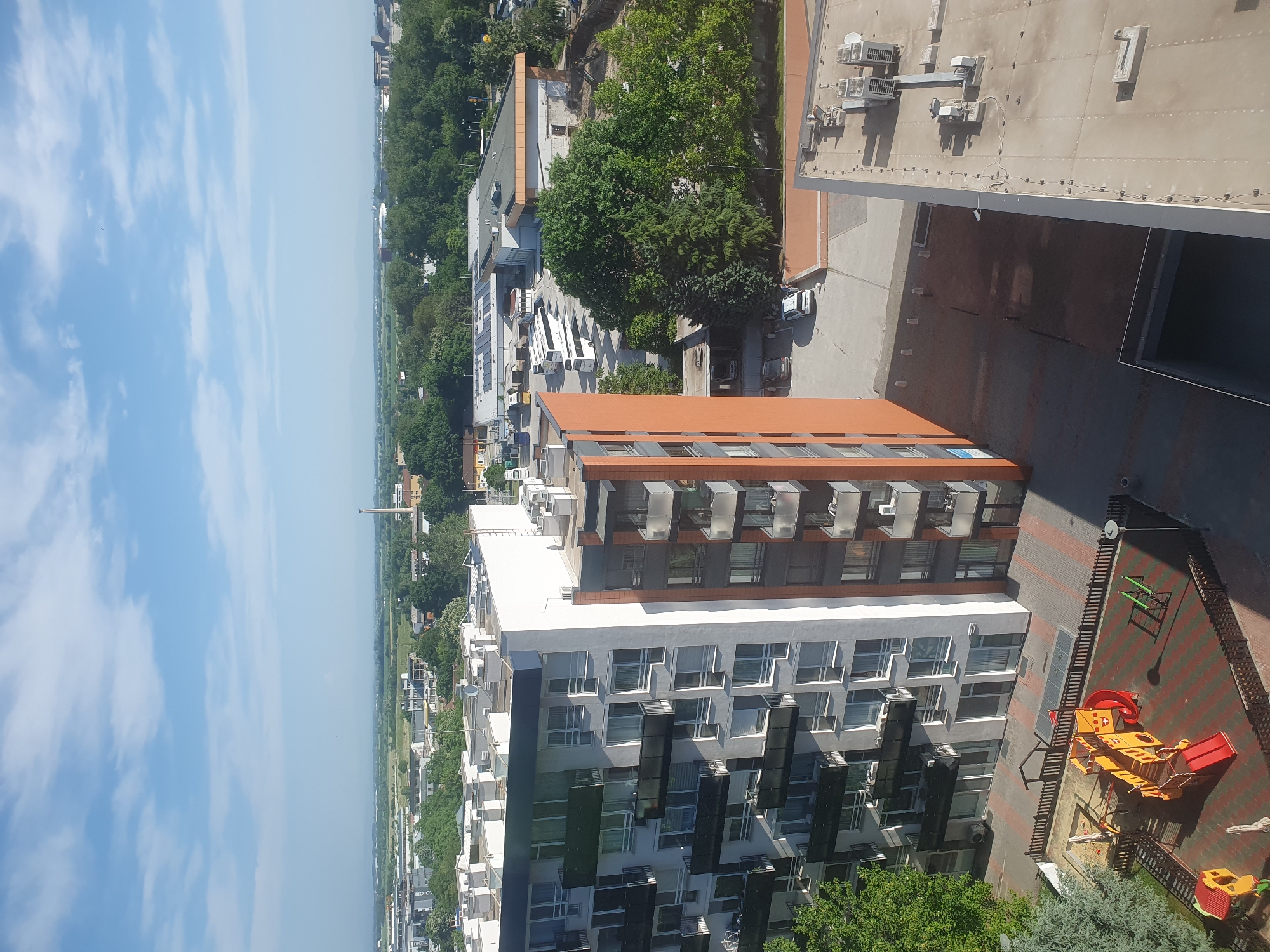 The image shows an elevated view of a residential area in Plovdiv, Bulgaria, featuring modern apartment buildings. The foreground is dominated by a tall building with a white and gray facade, adorned with balconies and dark panel accents. Adjacent to this building, there's a small, brightly colored playground with yellow and red play equipment. The backdrop is a mixture of urban and natural elements. In the middle ground, there are several buildings, including some with orange roofs, interspersed with trees and greenery. The horizon is visible, with a light blue sky containing scattered clouds. The overall impression is that of a contemporary urban setting with residential and recreational spaces.