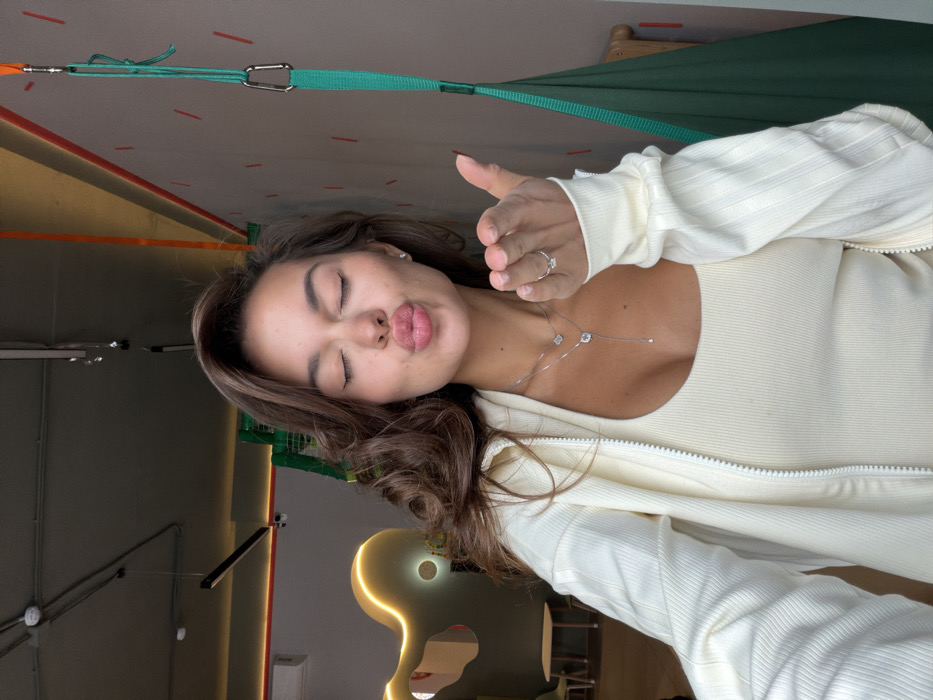 A woman poses in a casual, light-colored ribbed outfit, including a tank top and zip-up jacket. She purses her lips in a kissing gesture. A delicate silver necklace adorns her neck, and a ring is visible on her finger. The background includes room fixtures like ceiling beams, a light fixture, and a glimpse of what appears to be green netting. The photo has been rotated, altering the perception of perspective.