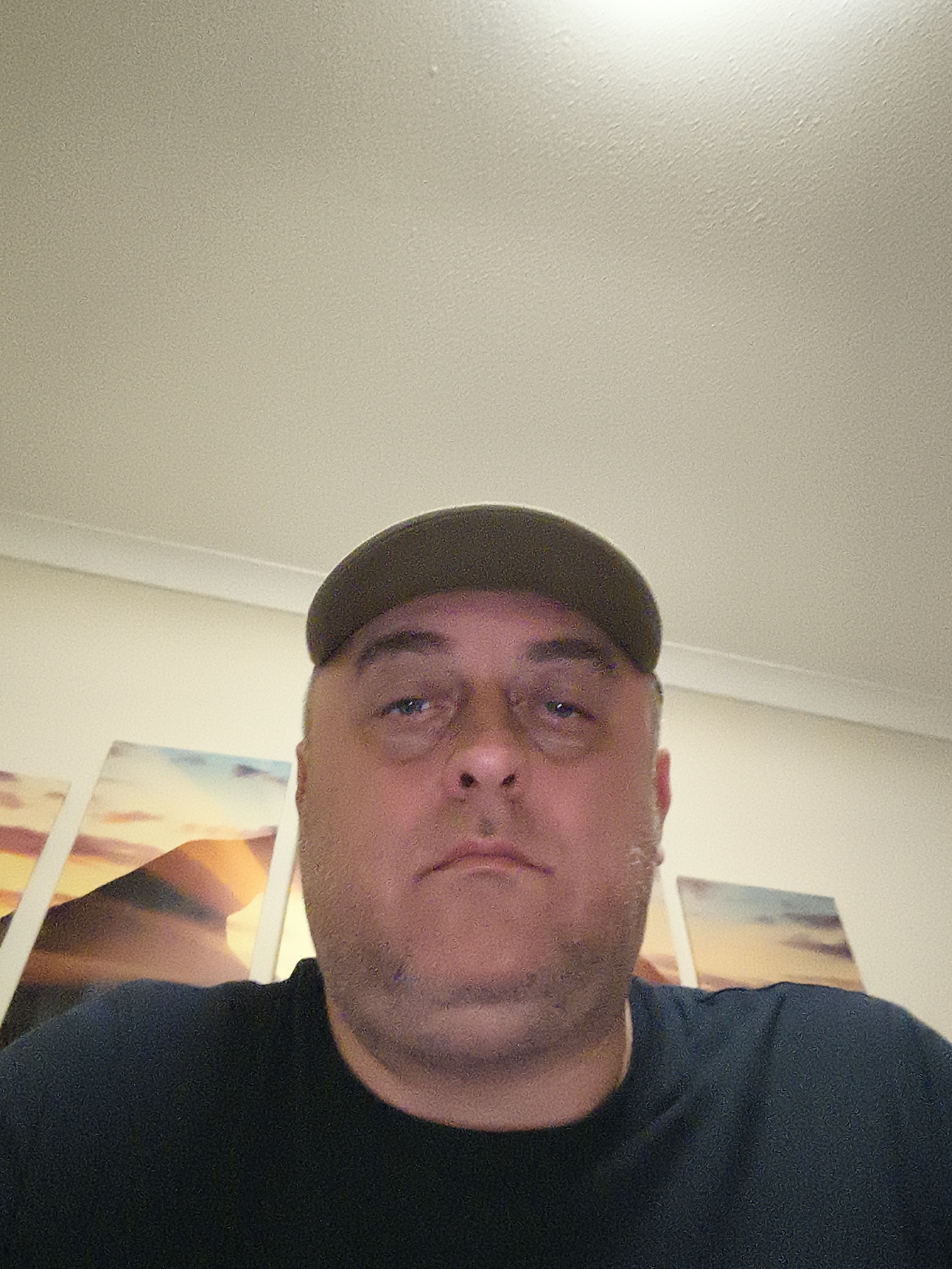 A portrait captures a man indoors, believed to be in Huntingdonshire, United Kingdom. He is positioned centrally, facing the camera directly, and appears to be in his middle age. He wears a dark olive green or khaki baseball cap and a plain dark crew-neck t-shirt. His expression is neutral, possibly conveying a sense of weariness, with a full face and visible stubble along his jawline.
Behind the man, on a light-colored wall, several pieces of art are displayed side-by-side. These appear to be abstract or landscape prints, featuring a blend of warm orange and yellow hues with cooler blue and purple tones, reminiscent of desert or sunset scenes. Above the man, a textured, light-colored ceiling is visible, illuminated by an overhead light source positioned towards the top center of the frame. The overall image has a somewhat grainy quality, indicating digital noise, possibly due to low light conditions or camera settings. There is no visible text.
