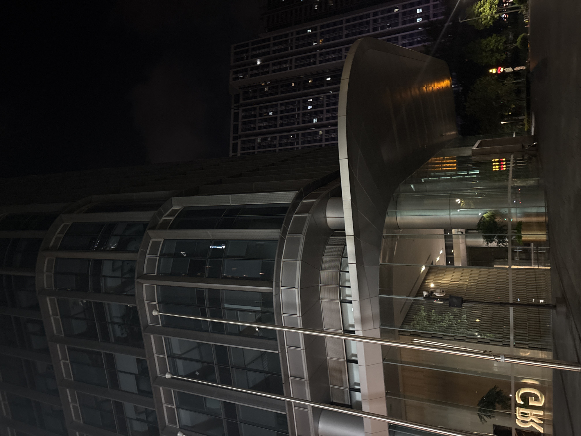 This is a nighttime, eye-level, low-angle shot of a modern building in Haizhu District, China. The building features a sleek, curved, metallic facade on the right side of the frame, contrasting with the geometric, rectilinear design of another building visible in the background. The larger building in the background has numerous windows, some of which are illuminated, suggesting occupancy. Reflections of the building's interior and the surrounding environment are visible on the glass surfaces.
The scene is illuminated by artificial lighting, indicating it is nighttime. The sky is dark, and there are no visible weather conditions. The architecture suggests an urban environment, likely a business or commercial district. The overall mood is modern and somewhat dramatic due to the lighting and angles.
The primary objects in focus are the architectural elements of the buildings. There are no discernible people or animals. The activity, if any, is implied by the lit windows.
There is visible text on the curved facade in the lower right corner, appearing as illuminated white characters on a dark background. The characters are stylized and appear to be in an East Asian script, possibly Chinese. They read "CEB".