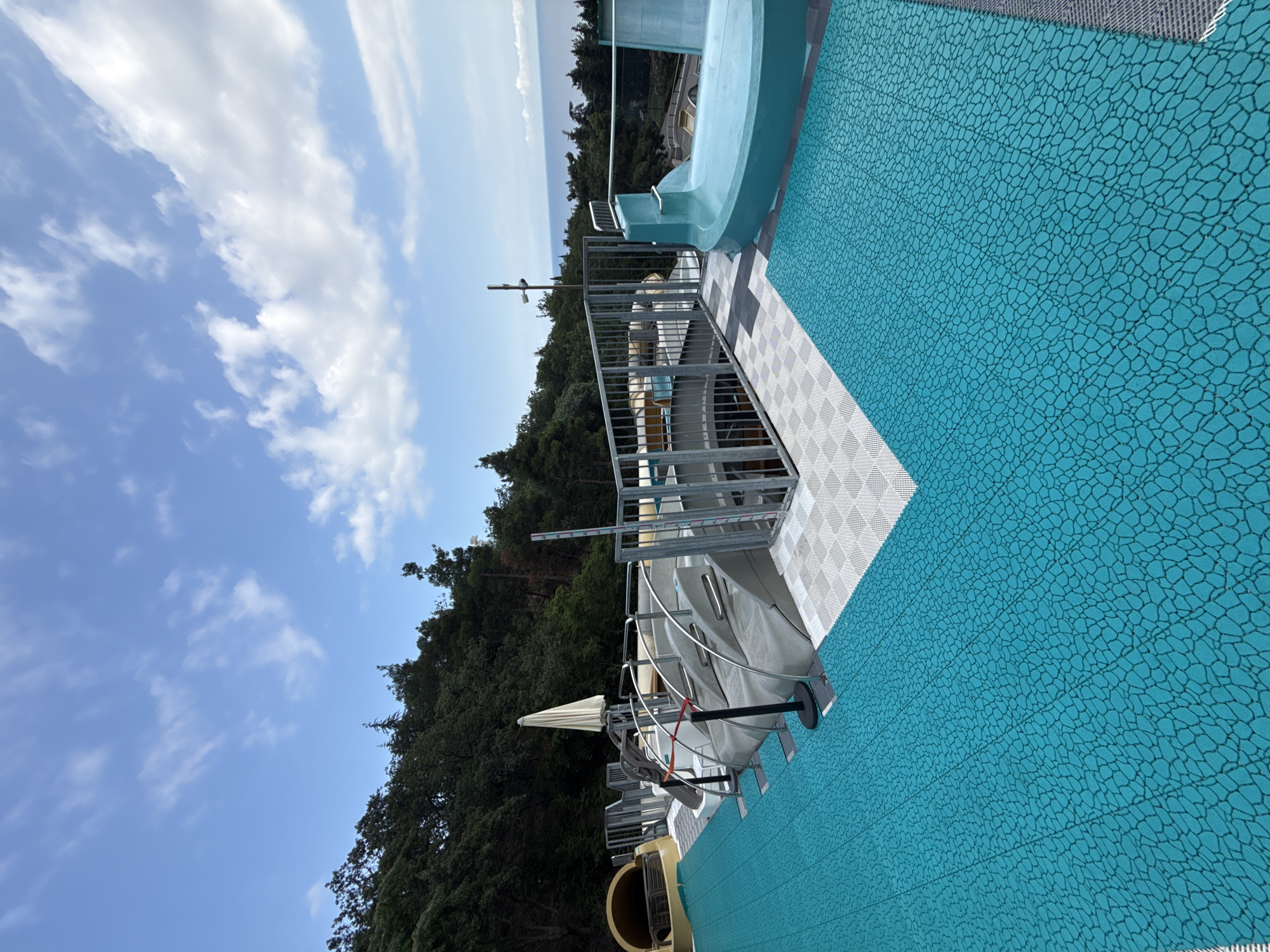 The image depicts a water slide complex situated outdoors with a view of the sea in the background. The foreground is dominated by a vibrant turquoise matting with a textured, irregular pattern, suggesting it's a non-slip surface for a pool or water park. Beyond this matting, there are tiered grey structures that appear to be the seating or waiting area for the water slides, complete with metal railings. Partially visible are portions of yellow and blue water slides. A closed, light-colored umbrella stands to the left. In the distance, dense green trees line a hillside, and the blue expanse of the sea meets the horizon under a sky dotted with white clouds. There are no people visible in the image. The setting appears to be a developed recreational area.