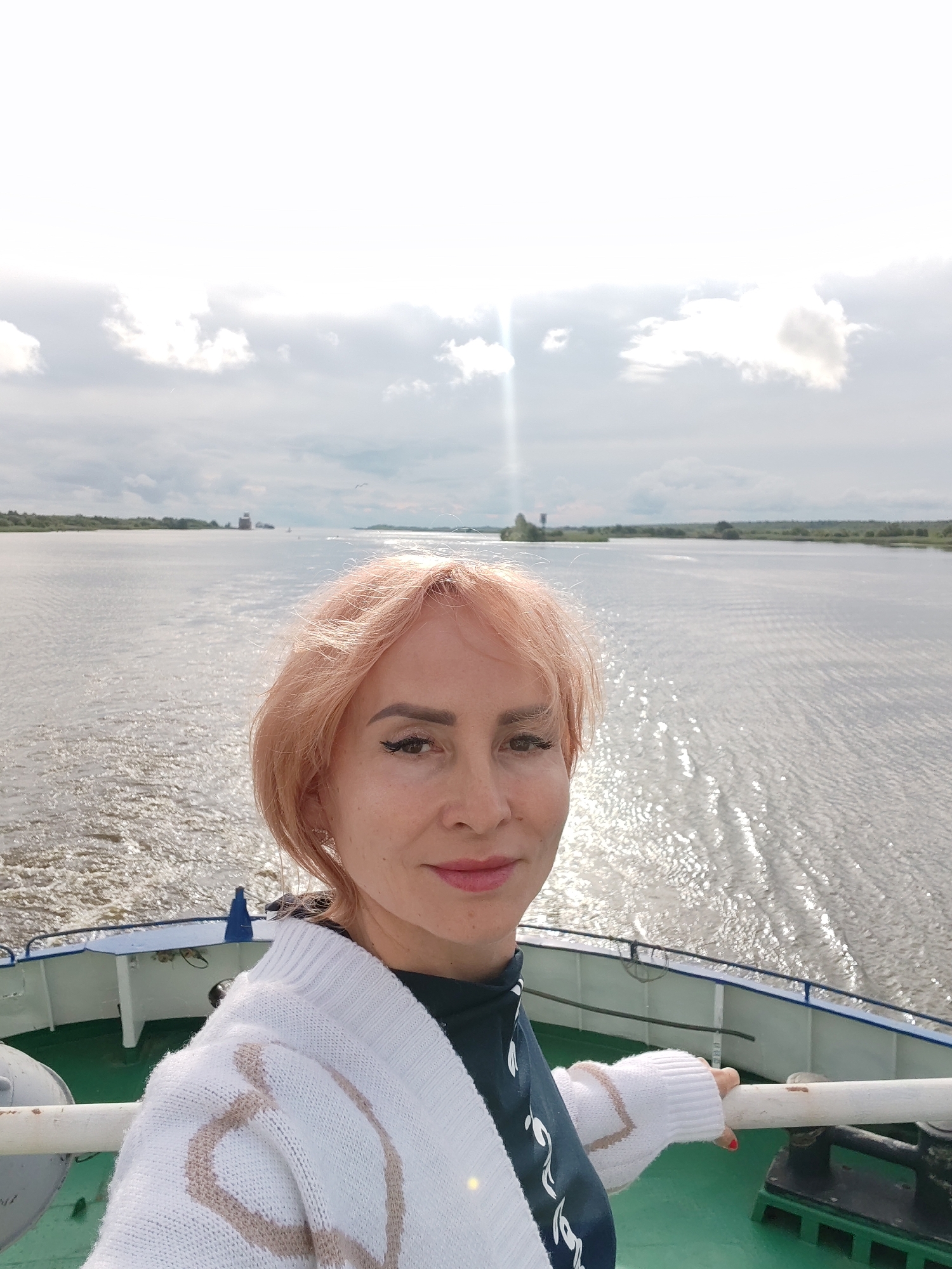 In the city of Kargulino, Russia, a fair-skinned woman with peachy-blonde hair takes a selfie on the deck of a ship. She is wearing a white cardigan with a tan pattern over a dark green top. Her right hand rests on the railing of the ship. The ship is on a body of water that appears to be a river, with a green deck visible. The water is rippled and reflects the light from the sky, which is overcast with some bright spots indicating the sun. In the background, there is a distant shoreline with trees and a large barge or ship.