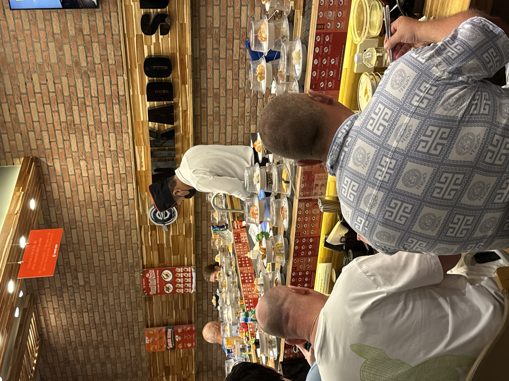 Here is a description of the image:
The image captures a scene in a Mikado sushi restaurant, located in Nha Trang ward, Vietnam. A conveyor belt of sushi rotates in front of several customers. The focus is on a group of diners: one in a Versace pattern shirt using chopsticks, another in a white shirt with a green design. In the background, a chef wearing a white uniform and a mask prepares food behind the sushi belt. Menu boards for "Hand Roll Sushi" and "Aburi Sushi" are visible on the brick walls, as is a sign stating food not on the conveyor belt can be ordered. The restaurant's name, "MIKADO," is displayed in large letters along the wooden wall.