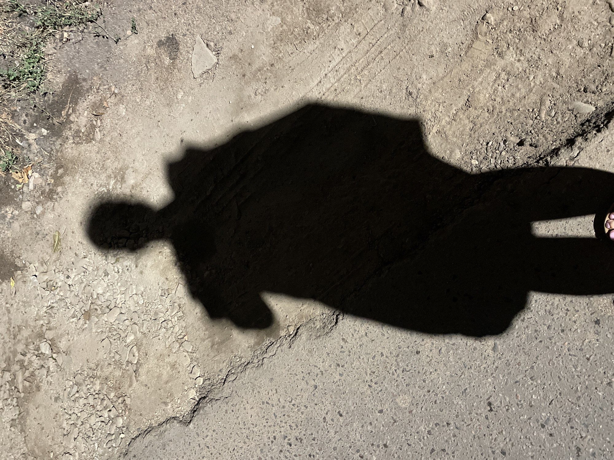 A shadow of a person holding an umbrella is cast on a cracked asphalt surface with gravel and dry grass bordering the path. The scene is an outdoor environment, possibly a street or sidewalk. The shadow indicates the presence of a person with an umbrella, standing in a position that suggests they are facing away from the camera. The ground texture varies between smooth asphalt, rough gravel, and sparse vegetation. The lighting suggests it is daytime, with the sun casting a strong shadow. There are no discernible landmarks in the image that would specifically place it in Almaty, Kazakhstan.