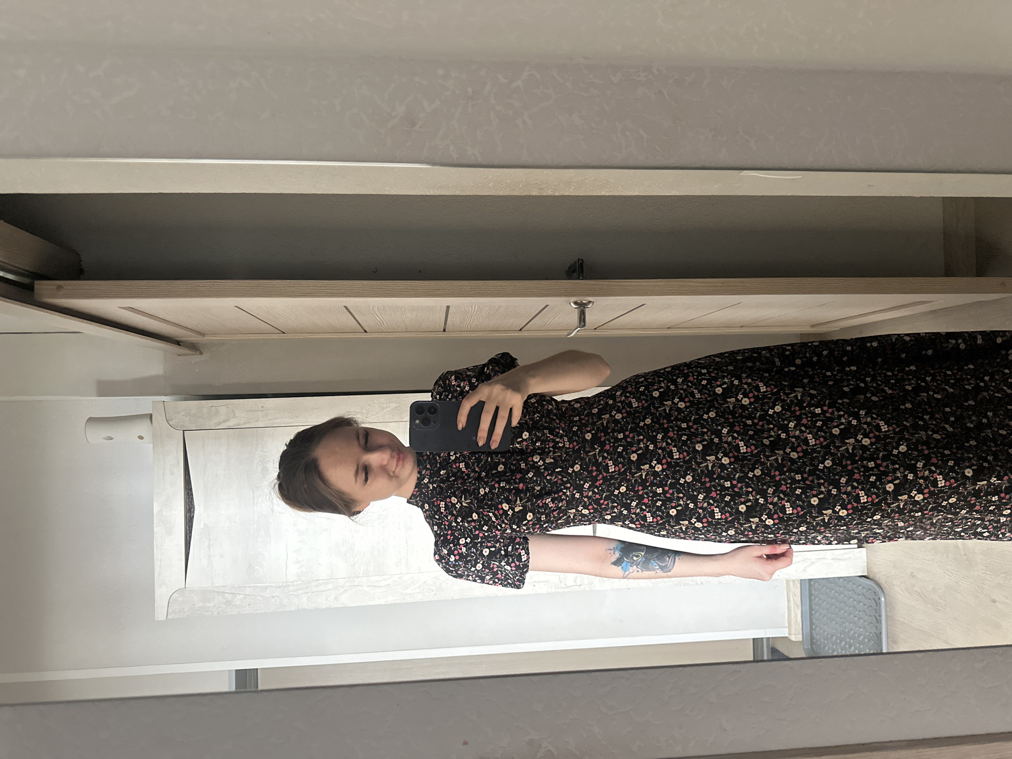 A female adult with light brown hair pulled into a bun is depicted taking a mirror selfie within what appears to be a private residence in the city of Unknown, Unknown. She is wearing a dark-colored, high-necked maxi dress with short puffed sleeves, adorned with a dense pattern of small pink, white, and possibly light green flowers. A distinct, colorful tattoo of a blue creature with large green eyes is visible on her left forearm. She holds a dark-colored smartphone in her right hand, aiming it towards the mirror, and is looking down at the phone's screen with a subtle, soft smile.
The mirrored background reveals light-colored walls, with some sections displaying a subtle textured pattern. The flooring appears to be light-toned wood or laminate. Two light-colored wooden doors are visible: one is a sliding door on the right side of the reflection, appearing partially open and revealing a darker space beyond, and another door with a silver handle is partially visible on the left side of the reflection. Above the door on the left, a small white wall-mounted shelf or cabinet is present, upon which rests a tall, slender white object, likely an automatic air freshener dispenser. A rectangular grey textured mat or tray is visible on the floor in the lower left portion of the reflection.