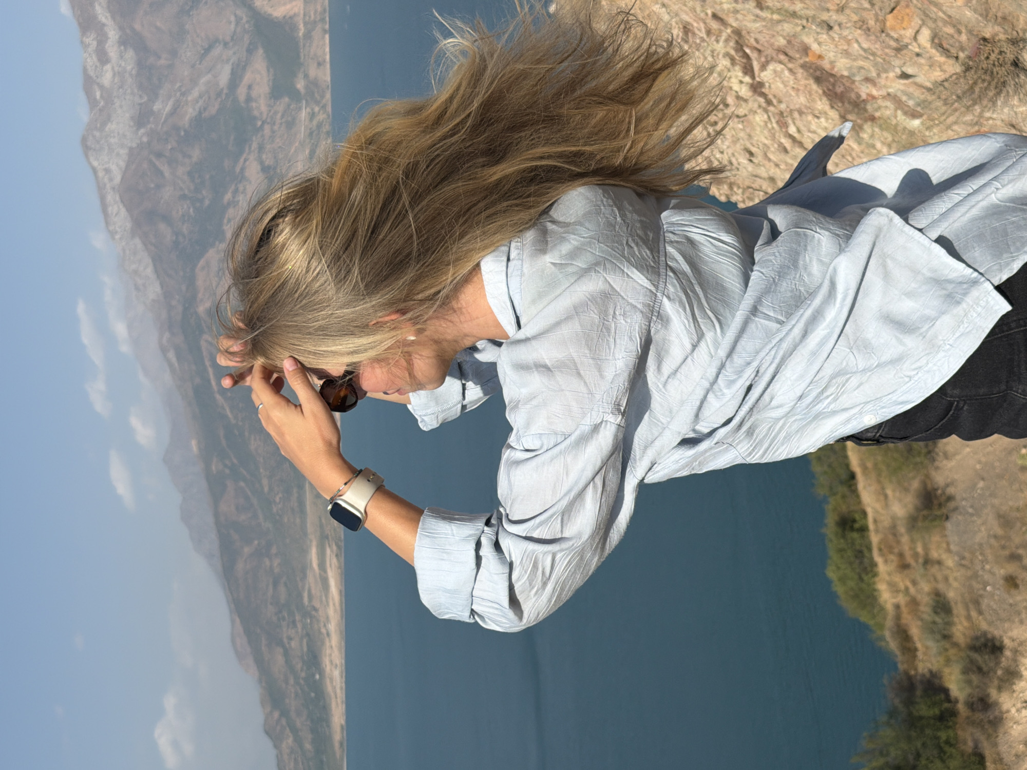 In the city of Chorvoq, Uzbekistan, a person stands on a rocky, arid hillside overlooking a large, calm body of water. The person, facing away from the camera and to the left, has long blonde hair that is blowing in the wind. They are wearing a light blue, crinkled button-down shirt and dark pants. On their left wrist, they are wearing a silver smartwatch. Their right hand is raised to their forehead, shielding their eyes from the sun, and their left hand is holding their hair out of their face. In the background, distant mountains rise under a clear blue sky.