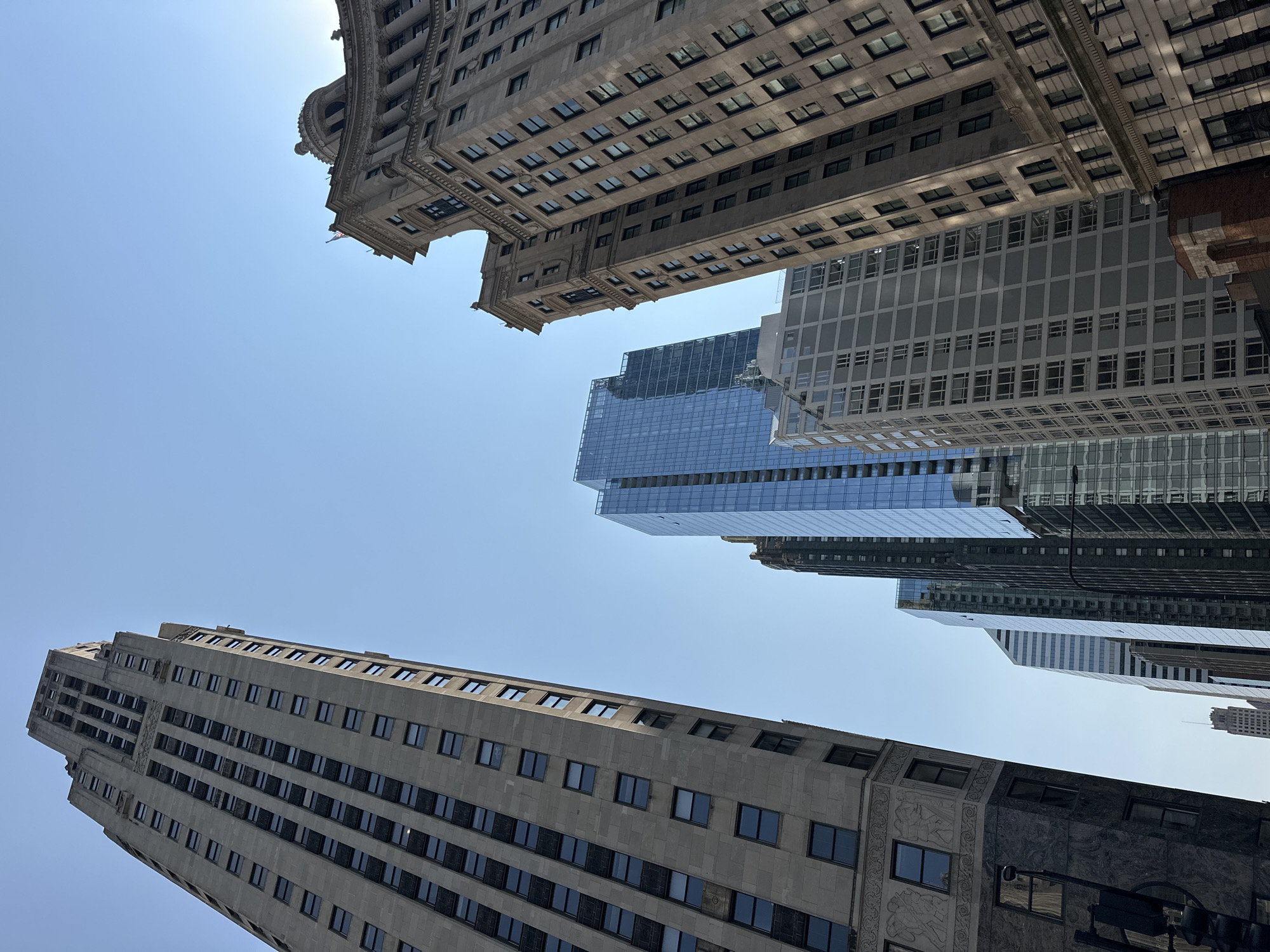 The image captures a low-angle perspective of several skyscrapers in Chicago, Illinois, USA, against a clear blue sky. The buildings are varied in architectural style and material. On the left, a tall, grey stone building with a distinct art deco design dominates the frame, its numerous windows reflecting the sunlight. To its right, a modern skyscraper with a glass facade is visible, its blue and silver tones contrasting with the older structures. Further to the right, a more ornate, classical building with intricate stone carvings and a domed roof is partially visible, also in stone. Other buildings, appearing to be of varying heights and designs, fill the background, creating a dense urban landscape. The sunlight casts distinct shadows on the buildings, indicating the time of day. No people are visible in the image. The overall scene is one of urban density and architectural diversity in a major metropolitan area.