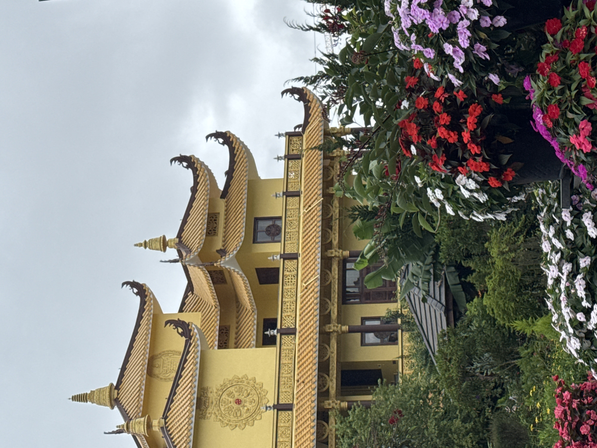 The image features a yellow, multi-tiered building in Da Lat, Vietnam. The building has intricate architectural details, including dragon-shaped roof ornaments and patterned exterior walls. The sky is overcast. In the foreground, a variety of flowers in red, white, and purple hues add color to the scene, partially obscuring the building. Lush greenery surrounds the building, blending with the flowers to create a natural environment.