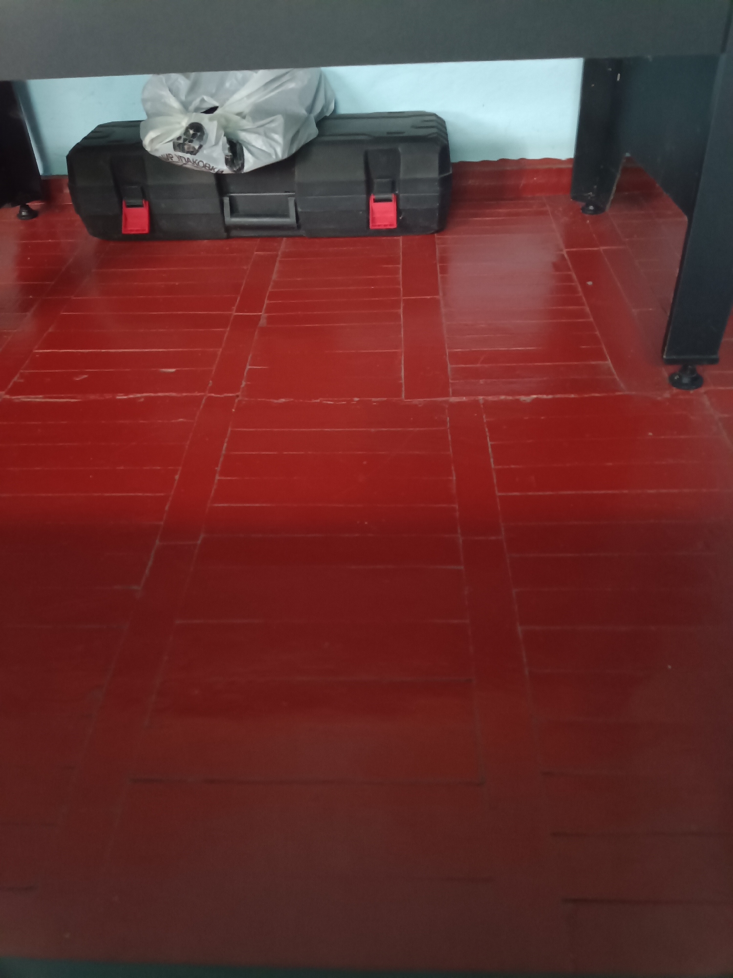 A black toolbox with red clasps and a white plastic bag on top sits on a red tiled floor beneath a table or desk. The floor is composed of rectangular tiles arranged in a grid pattern, with darker grout lines. The wall behind the toolbox is light blue. Part of the dark legs of the table or desk are visible on the right side of the image. The scene is indoors, in a room with a flat, light blue wall and a red tiled floor. The location is the city of Kenzhe, Russia.