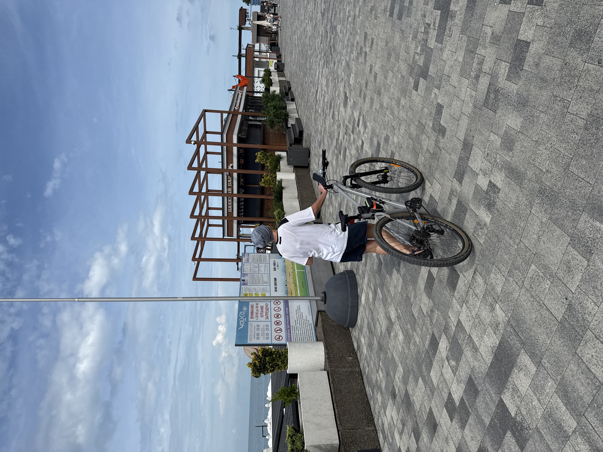 A person with a bicycle walks on a paved promenade in Sochi, Russia, on a cloudy day. The person, wearing a gray cap, white t-shirt, and dark shorts, is seen from behind, pushing a silver bicycle. The promenade is paved with gray and dark gray tiles arranged in a geometric pattern. On the left side of the promenade, there are white rectangular planters with greenery and a tall pole with a sign. In the background, there are beach chairs, umbrellas, and a row of beachside establishments, including a bar with a sign that reads "ПЛЯЖНЫЙ БАР" (Beach Bar). The sea is visible in the distance.