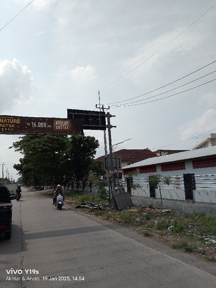 The image presents a daytime street scene in Arjawinangun, Indonesia, under an overcast sky. A paved road occupies the lower left and central portions of the frame, showing a dark-colored vehicle's rear on the far left and two individuals riding motorcycles further down the road. The road is flanked by green trees on the left and a building complex on the right.
On the right side of the road, there is a low building with a white wall and visible red brick accents, topped with a tiled roof. A modern-looking fence with black pillars and silver horizontal bars encloses the property. The roadside verge on the right appears somewhat overgrown and contains scattered debris, including corrugated metal sheets. Several utility poles with a network of overhead power lines are visible across the sky, with one prominent, taller pole supporting a large, dark brown sign that spans across the road.
The sign displays text including "NATURE", "RETEK" (likely "KRETEK"), "RP 16.000 exs", and "REVOLUSI KRETEK", along with a star rating. The bottom left corner of the image contains a watermark that reads "vivo Y19s Akhtar & Arvan 19 Jan 2025, 14.54", indicating the photo was taken at 2:54 PM on January 19, 2025. The scene captures typical daily activity, with people commuting on motorcycles in a semi-urban or suburban setting.
